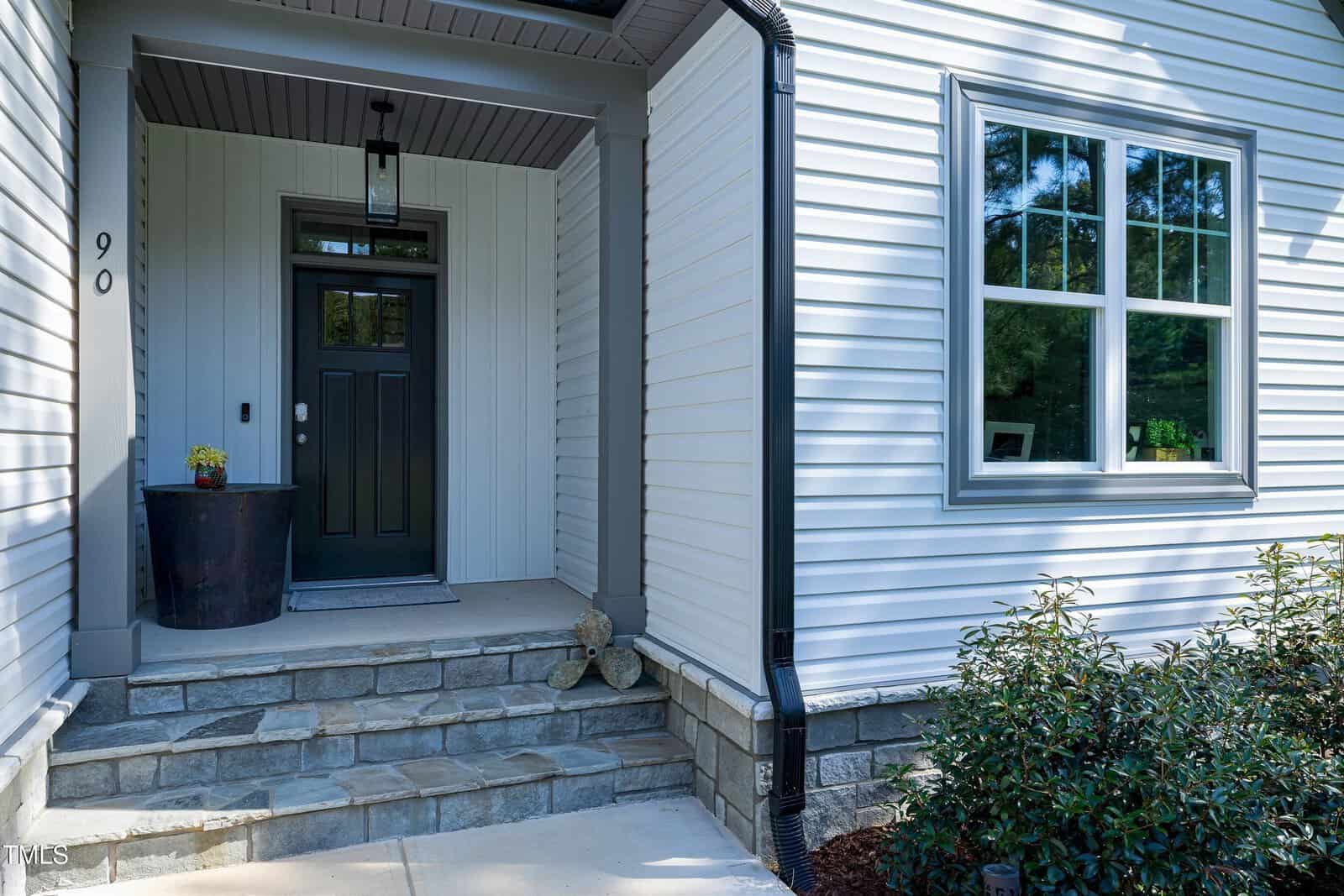 A modern house entrance with white siding, a black door, potted plant, stone steps, and a turtle statue on the porch. Large windows are on the right, with bushes in the foreground. House number 90 is visible.
