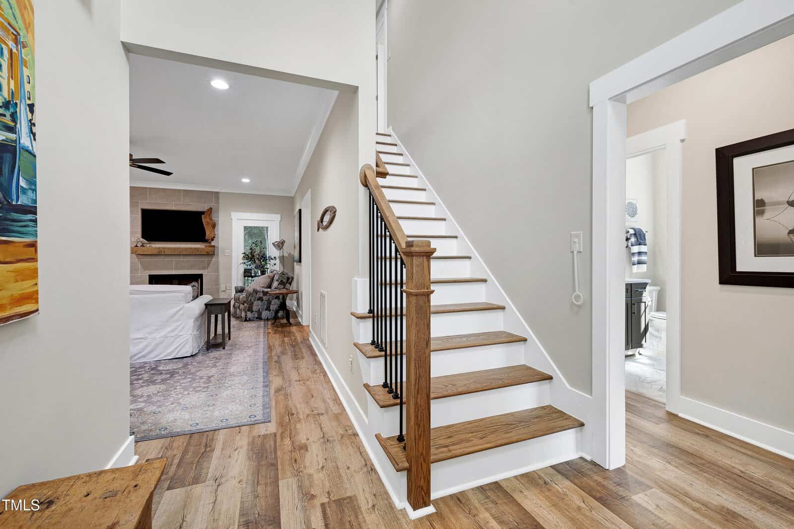 A bright hallway with wood flooring features a wooden staircase with white risers, leading upstairs. The living room with a fireplace and a white sofa is visible to the left, and a bathroom is seen through a doorway on the right.
