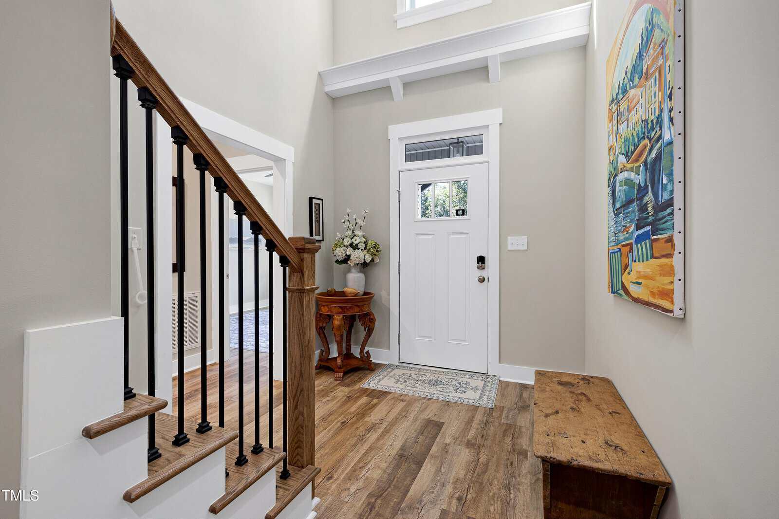 Bright entryway with a white front door, wooden floors, a staircase with dark railings, an artwork on the wall, a small table with a flower arrangement, and a rustic wooden bench. Natural light fills the space.