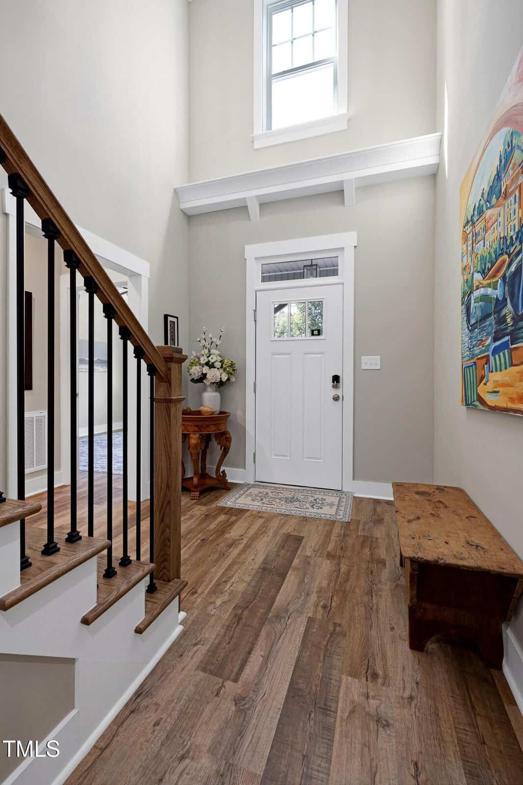 Bright entryway with wood floors, a white front door, a high window, staircase with black railings, wooden bench, floral arrangement on a small table, and colorful artwork on the wall.
