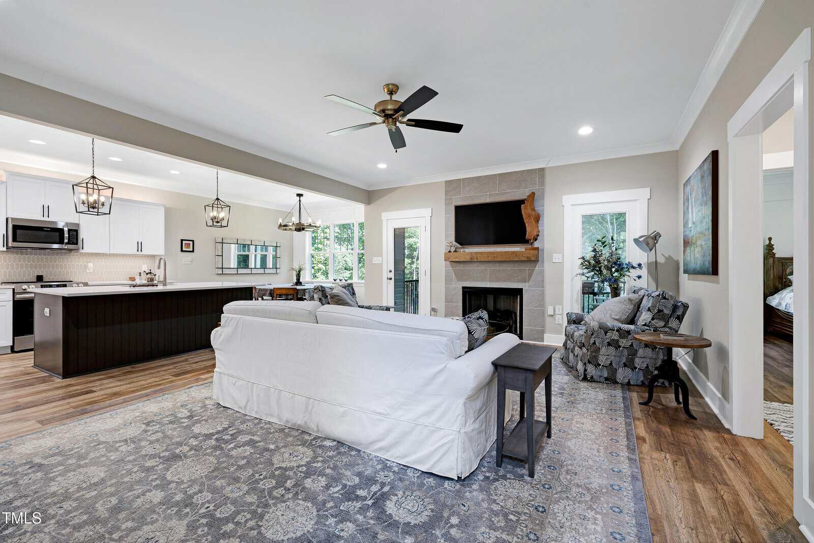 Open-concept living room and kitchen with white cabinets, stainless steel appliances, ceiling fan, stone fireplace with TV, gray sofa, patterned armchair, and large rug on wood floors. Natural light fills the space.