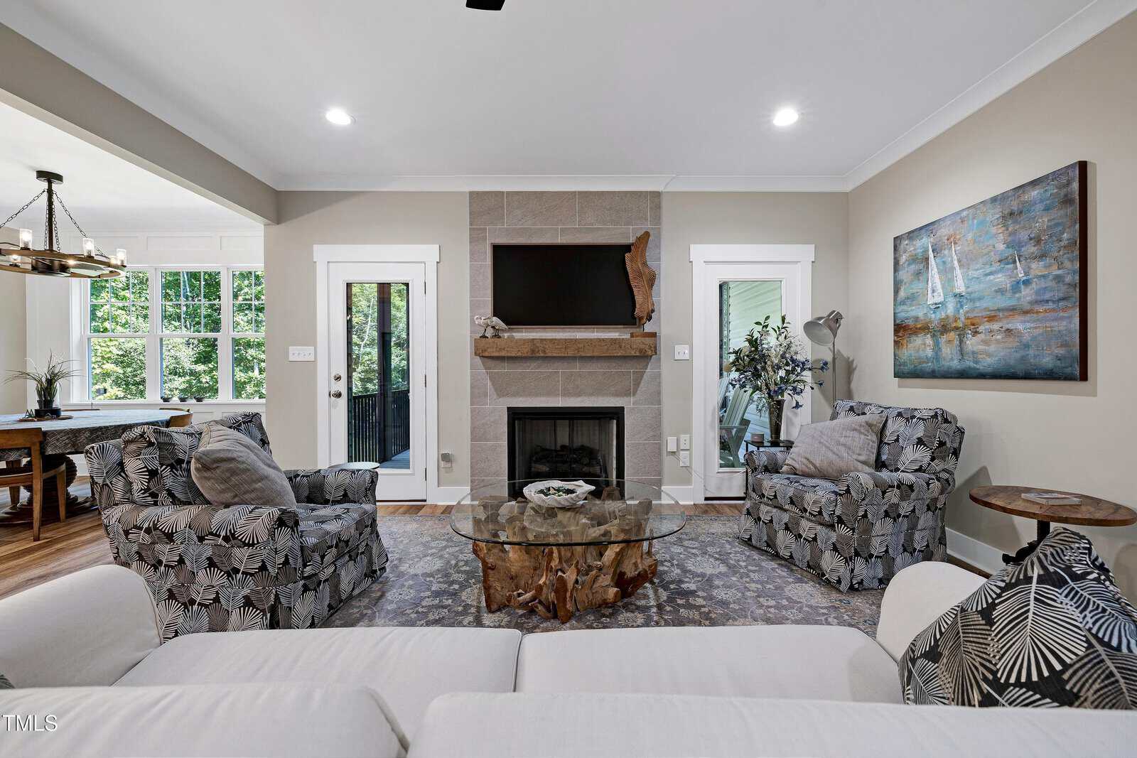 A cozy living room with two patterned armchairs, a white sofa, and a glass coffee table. A fireplace with a TV above it is centered on the back wall, flanked by doors and a large sailboat painting. Natural light fills the space.