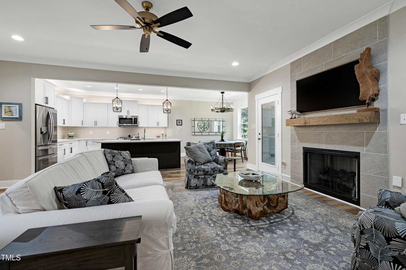 Open-concept living room and kitchen with modern decor, ceiling fan, white sofa, glass coffee table with a wood base, fireplace with mounted TV, and large windows allowing natural light.