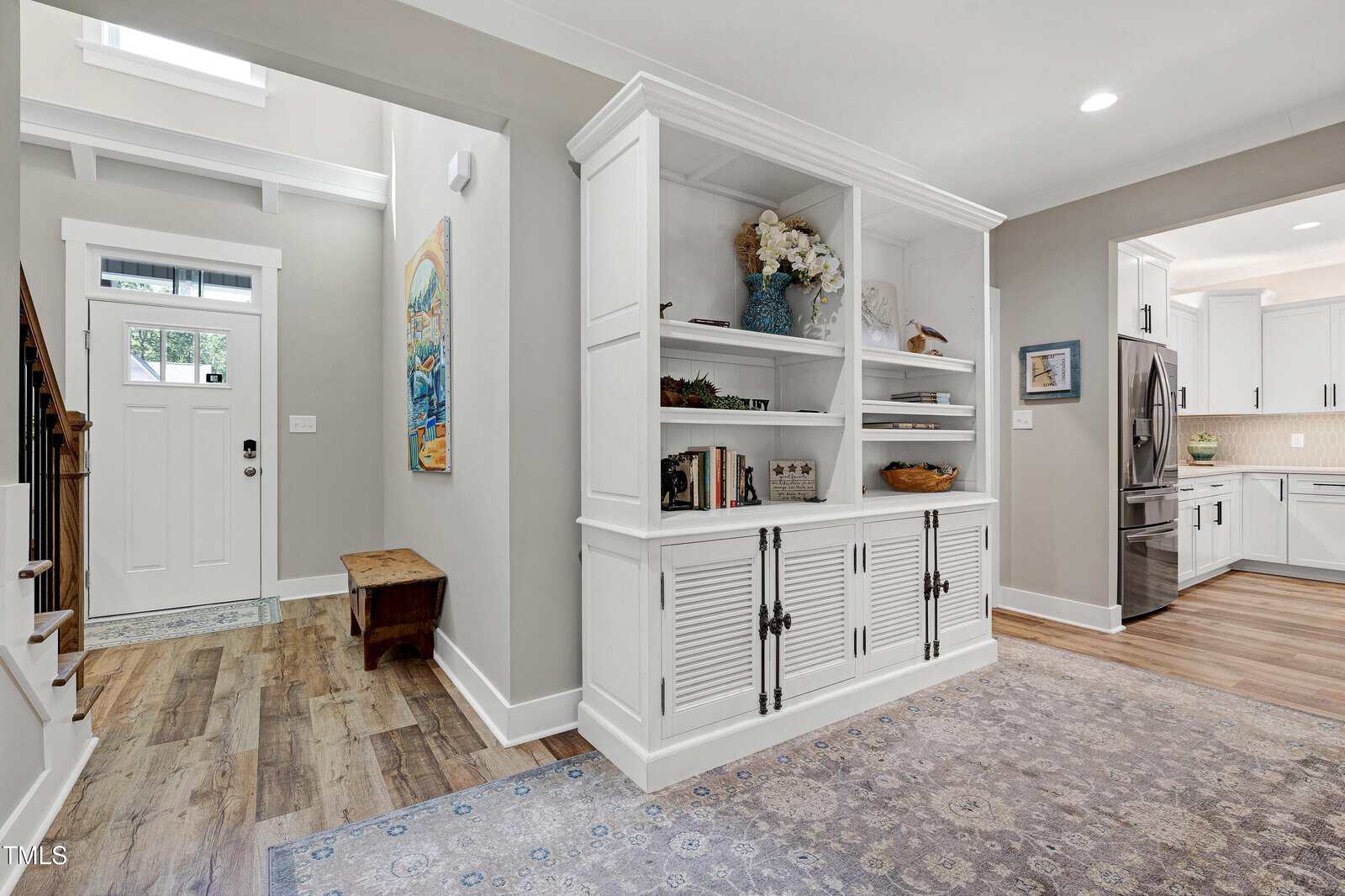 Bright entryway with wood floors, a white front door, a small bench, and a hallway leading to a kitchen. Built-in white shelves with decor and books divide the entry and living area. A patterned rug covers part of the floor.