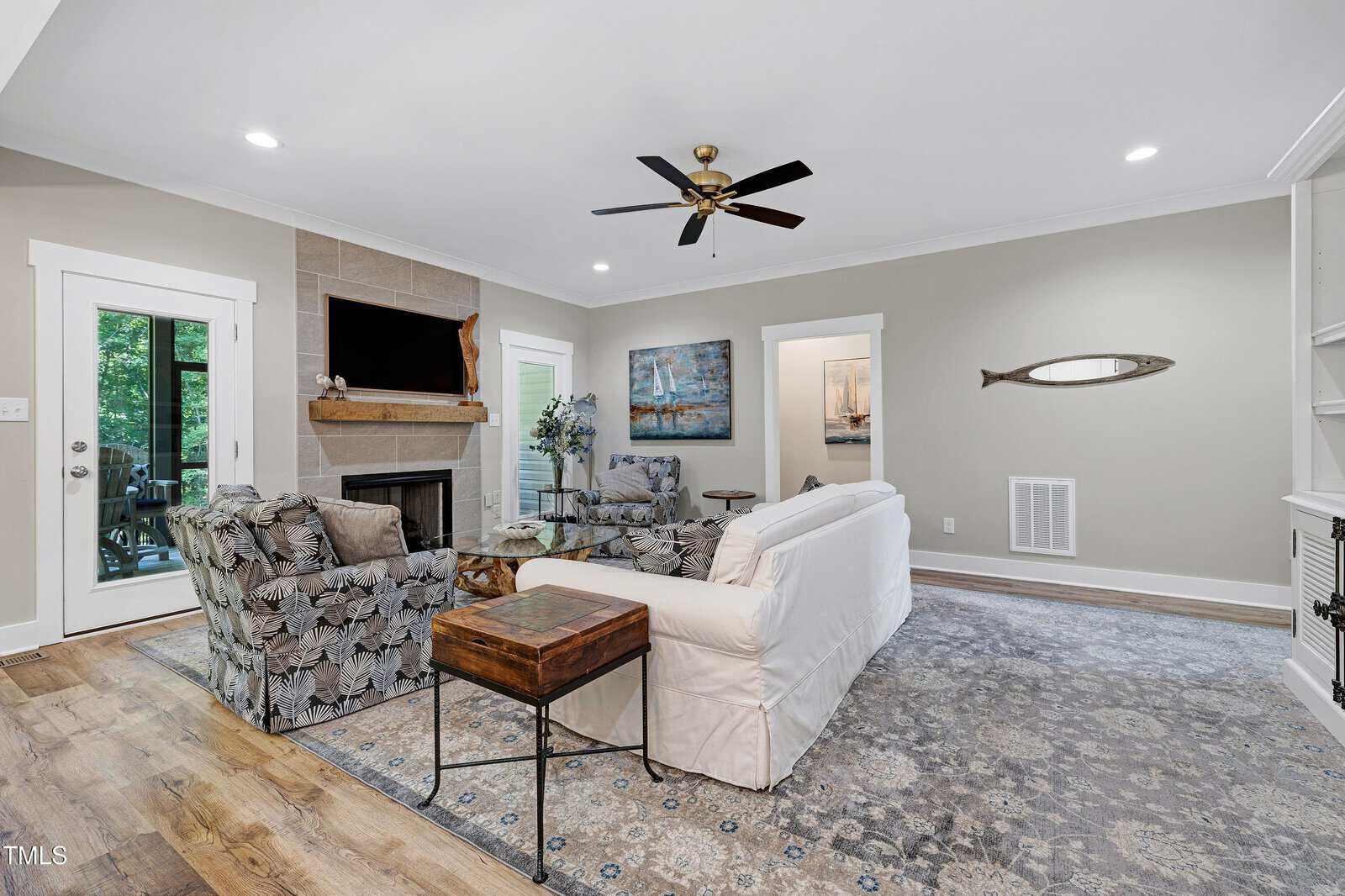 Bright living room with a white sofa, floral armchairs, rug, wall art, fireplace with TV above, ceiling fan, and built-in shelves. Hardwood floors and a glass door lead to the outside.