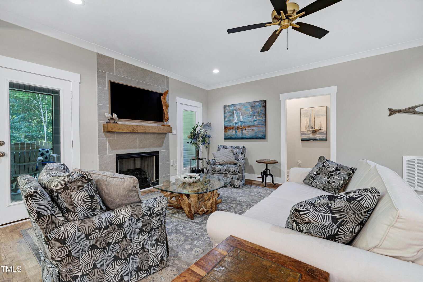 Cozy living room with patterned armchairs, a white sofa, fireplace with TV above, nautical wall art, ceiling fan, wooden coffee table, and door leading outside.