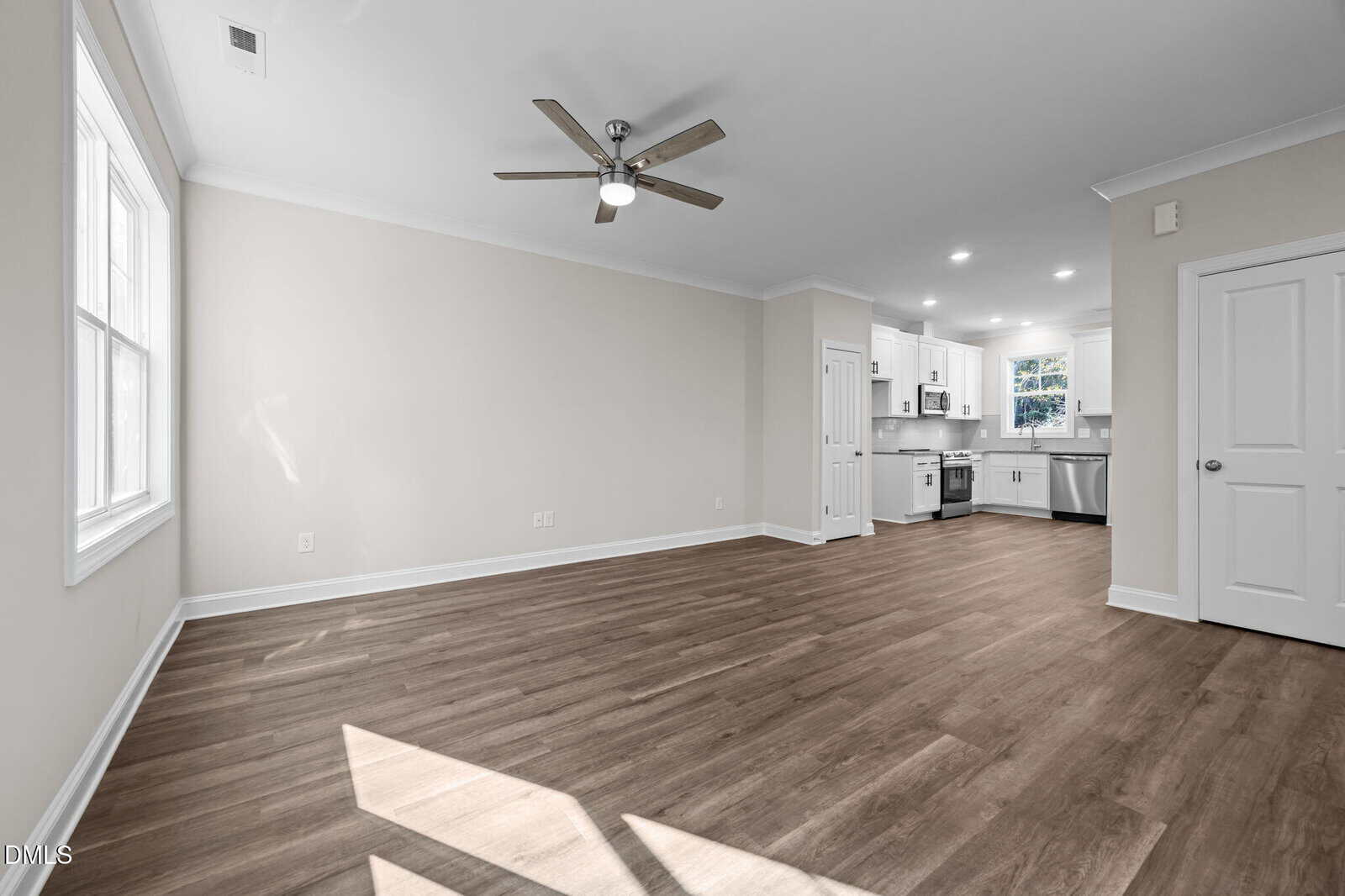 Bright, modern living area with wood flooring, large window, ceiling fan, white walls, and open access to a kitchen with white cabinets and stainless steel appliances.
