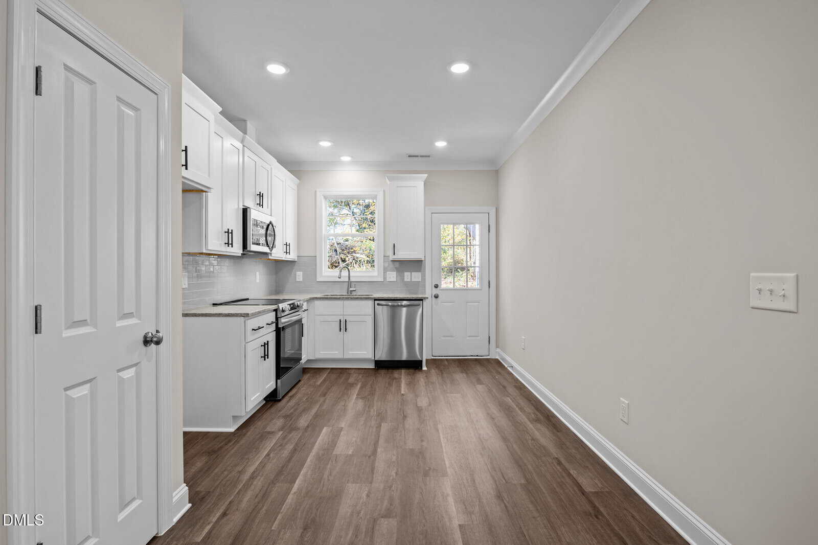 A modern kitchen with white cabinets, stainless steel appliances, and wood floors. A window and glass door let in natural light. The walls are painted a light neutral color, and recessed ceiling lights brighten the space.