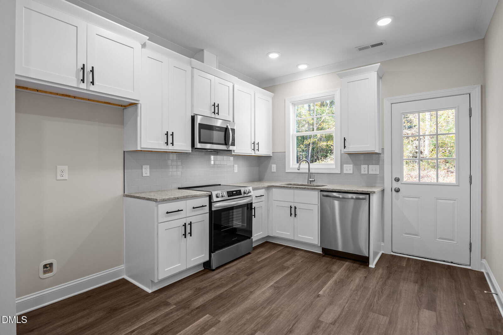 Modern kitchen with white cabinets, stainless steel appliances, a large window above the sink, light gray tile backsplash, wood-style flooring, and a white door leading outside.