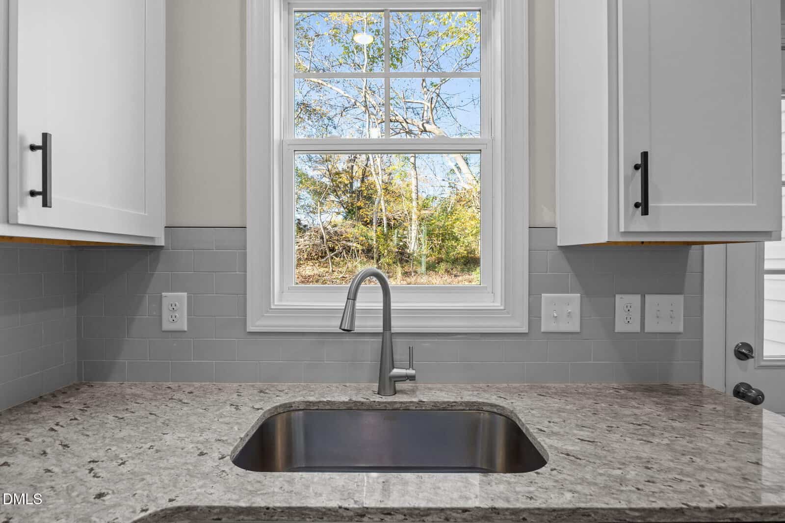 A modern kitchen sink with a granite countertop and stainless steel faucet is centered below a window, which shows trees and blue sky outside. White cabinets and light gray subway tile backsplash surround the sink area.