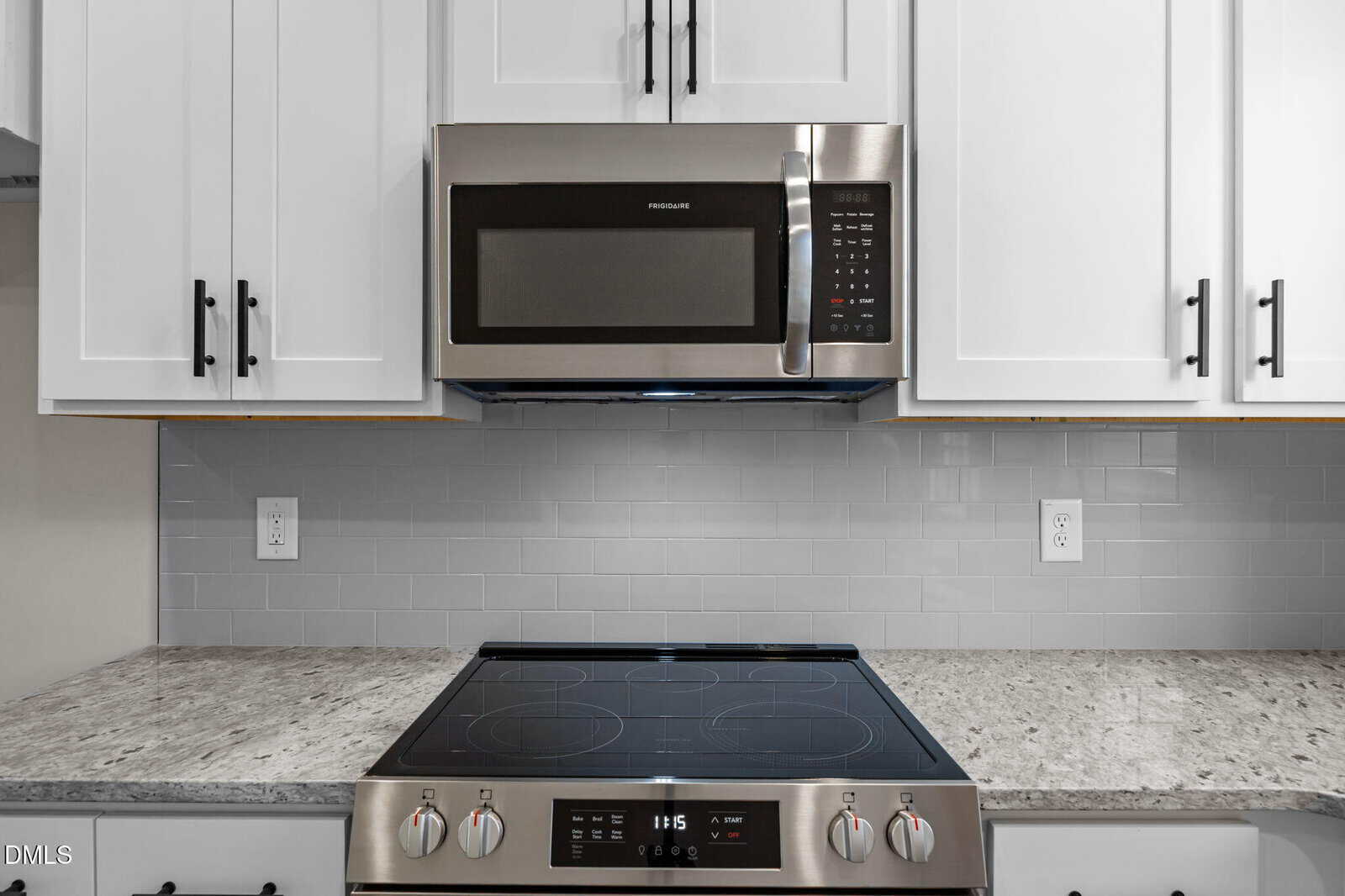 A modern kitchen features a stainless steel microwave above a matching electric stovetop, set against a glossy light gray subway tile backsplash and white cabinets with black handles.
