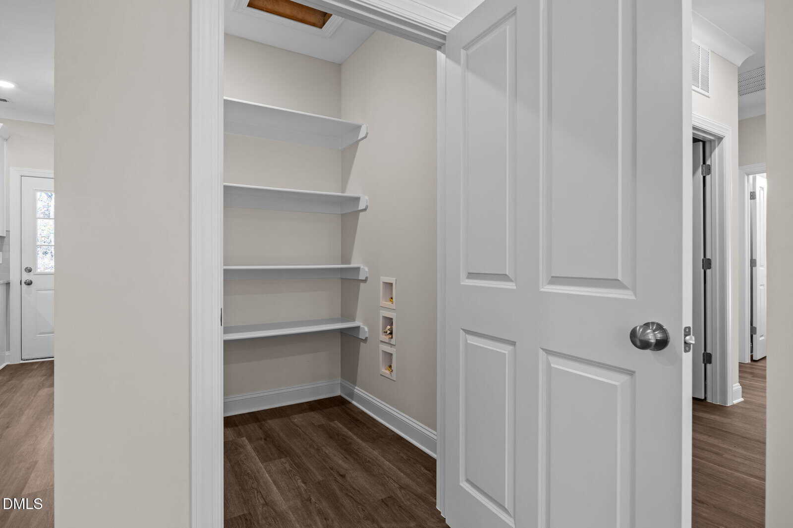 A small walk-in pantry with white shelves, wood-look flooring, beige walls, and an open white door. The pantry has three shelves and outlets on the side wall; a hallway with more doors is visible outside.