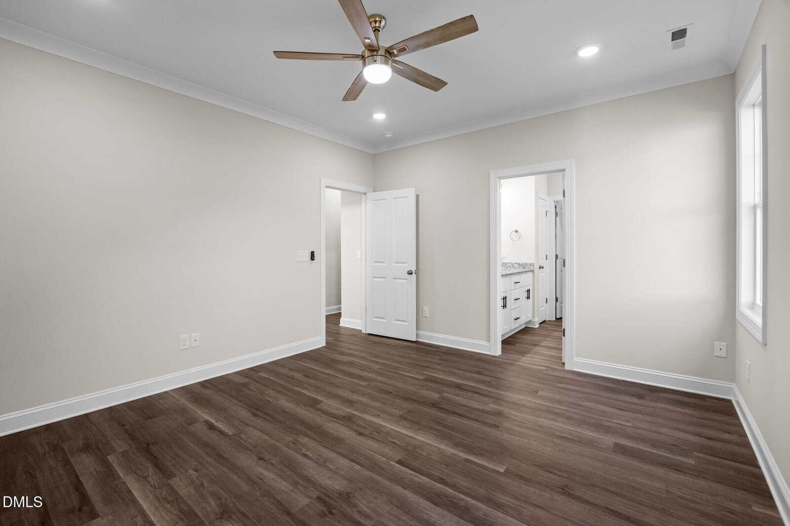Empty bedroom with light beige walls, dark wood flooring, a ceiling fan with light, white trim, and two doors—one leading to a bathroom with double sinks and the other a closet. Two windows provide natural light.