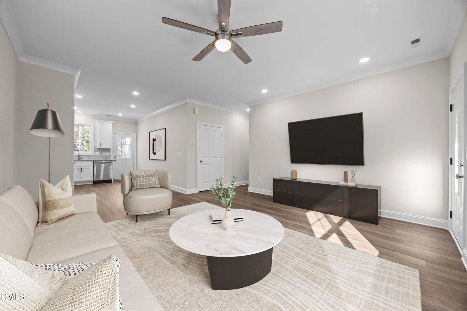 A modern living room with light walls, wood flooring, a beige sofa, round coffee table, armchair, floor lamp, large wall-mounted TV, and minimalistic decor. The kitchen is visible in the background.