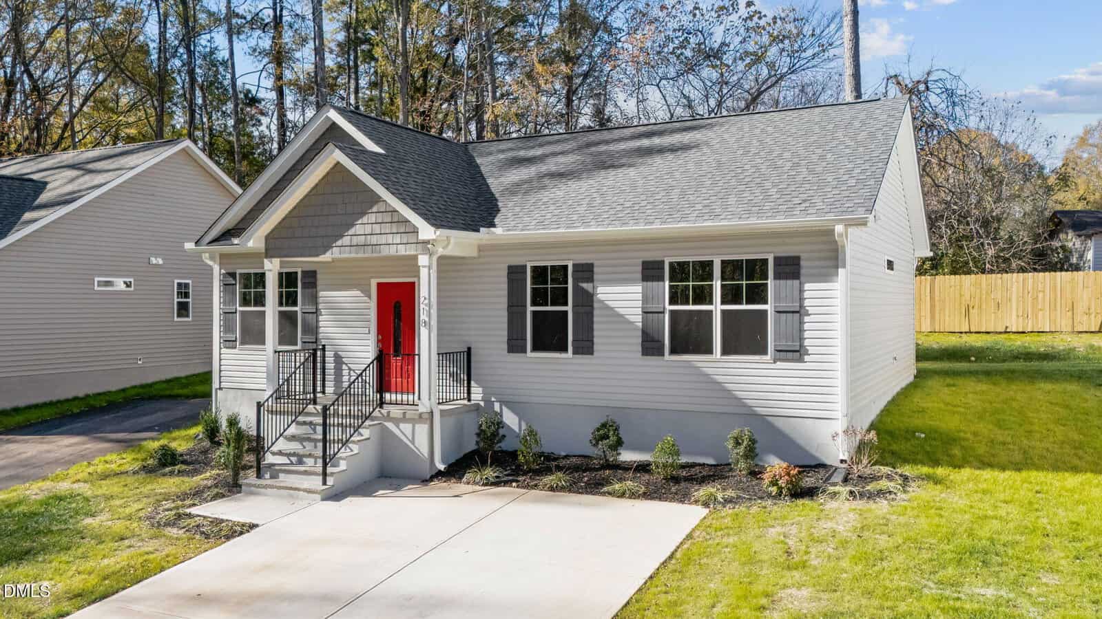 Single-story gray house with a red front door, black railings, small porch, landscaped front yard, and concrete driveway, set against a backdrop of trees and a wooden fence.