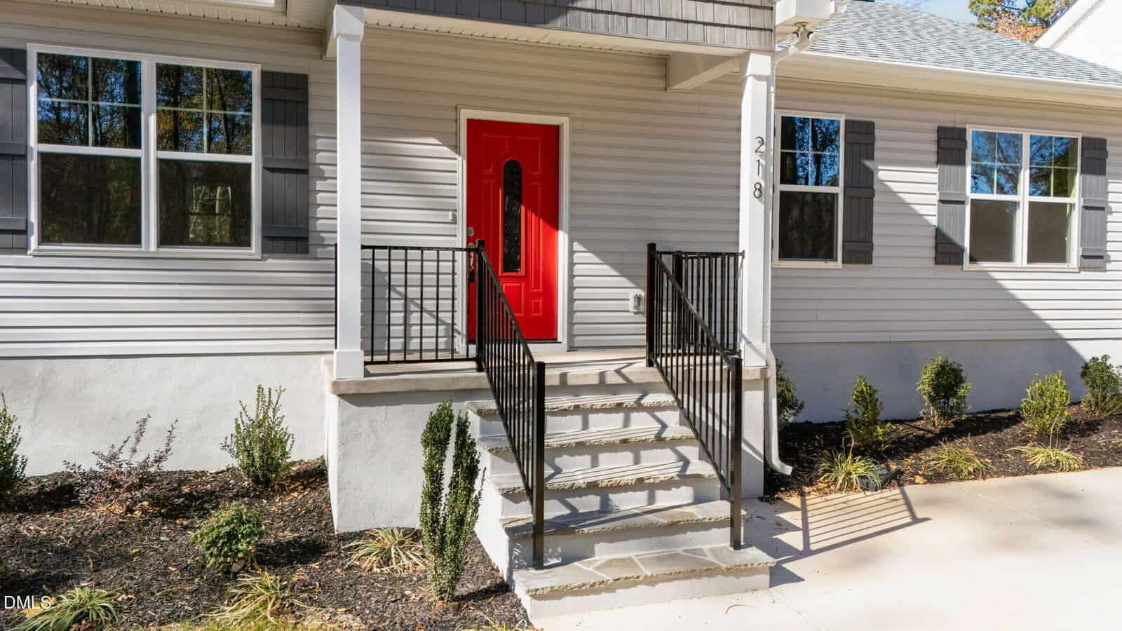 A gray single-story house with a bright red front door, white trim, and black railings on steps leading to the entrance. Small shrubs and plants line the walkway and house exterior.