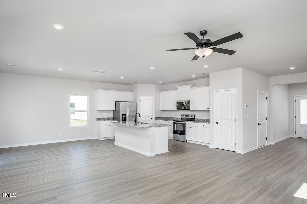 Open-concept kitchen and living area with light wood flooring, white cabinets, stainless steel appliances, a center island, and a black ceiling fan. Bright natural light comes in from a window.