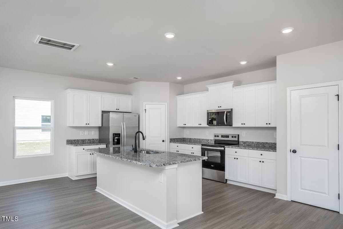 Modern kitchen with white cabinets, stainless steel appliances, granite countertops, an island with a black faucet, recessed lighting, wood-look flooring, and a window providing natural light.
