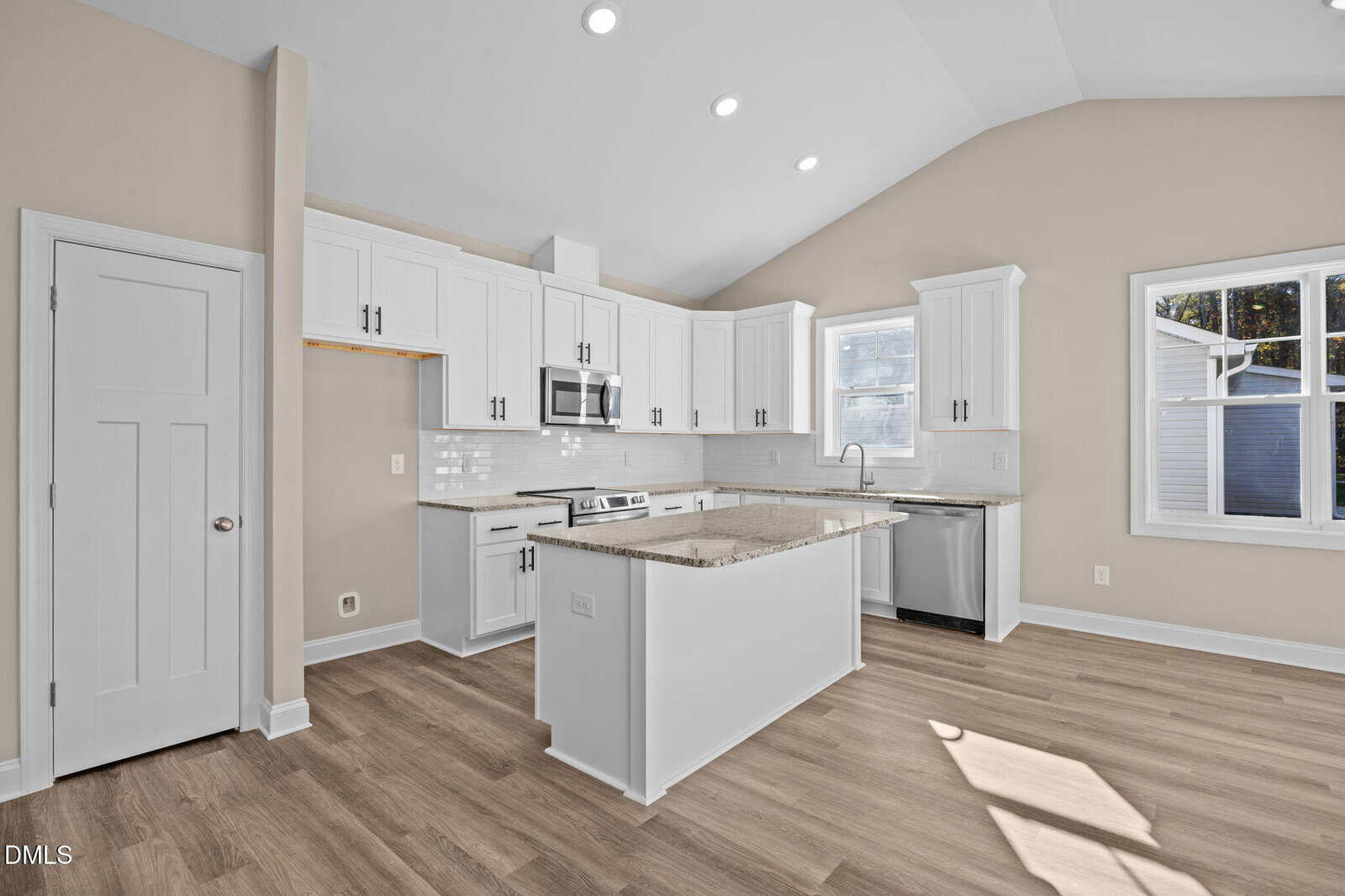 Modern kitchen with white cabinets, stainless steel appliances, a center island with granite countertop, light wood flooring, neutral beige walls, and large windows letting in natural light.