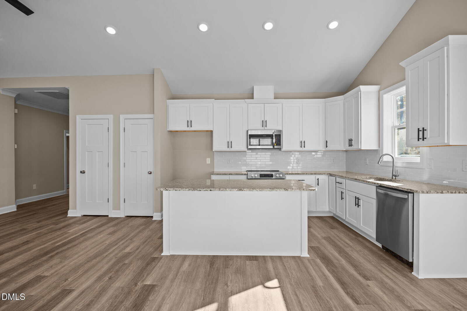 Modern kitchen with white cabinets, stainless steel appliances, an island with a granite countertop, light wood flooring, and neutral beige walls, illuminated by recessed ceiling lights and natural light from a window.