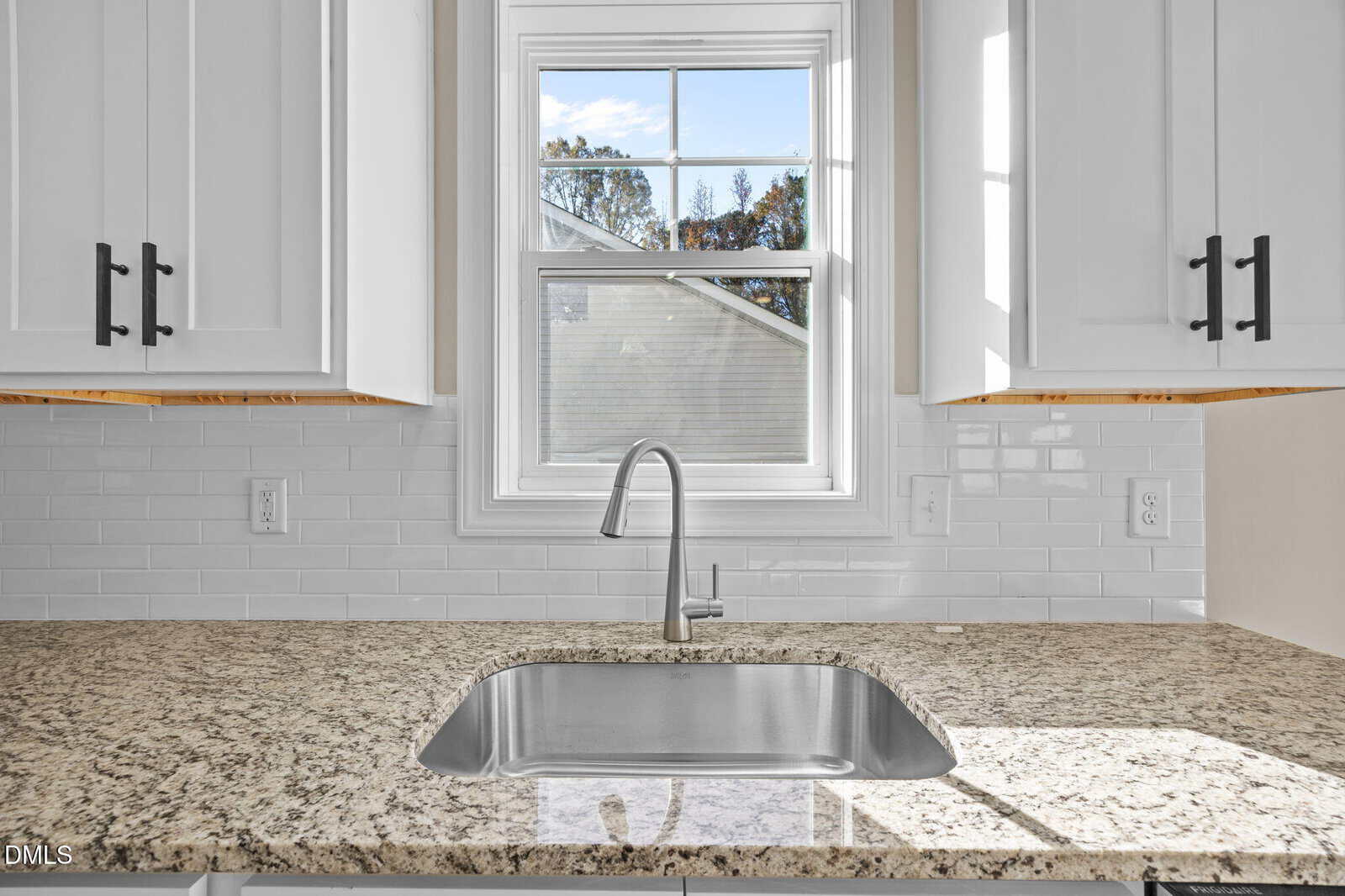 A modern kitchen sink with a chrome faucet set in a granite countertop, beneath a window with white trim. White tile backsplash and white cabinets with black handles surround the sink area.