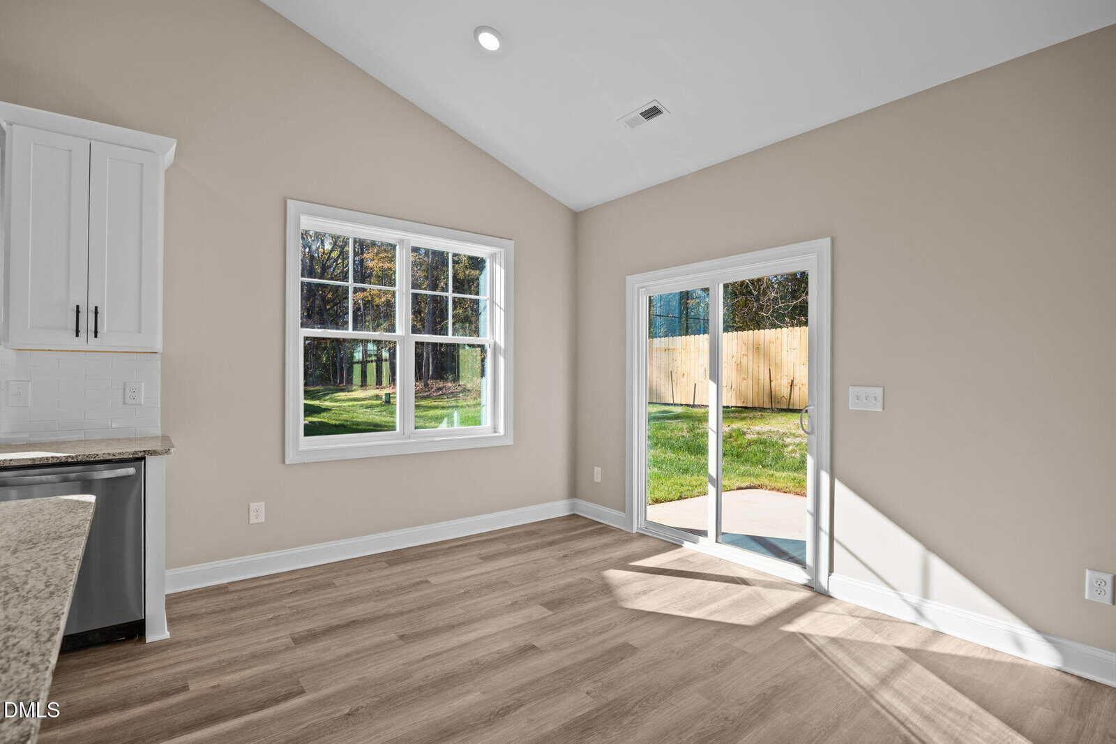 A bright, empty room with light wood flooring, beige walls, a large window, and glass sliding doors leading to a grassy backyard with a wooden fence. Sunlight streams through the doors, illuminating the space.