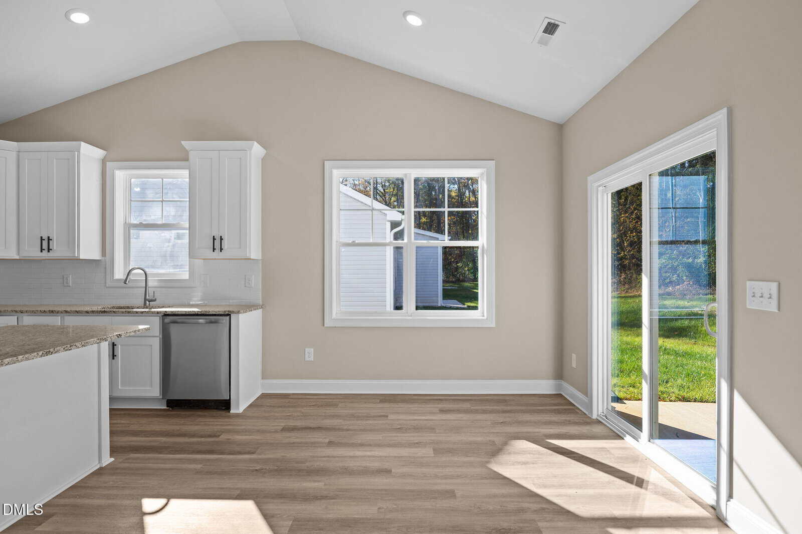 Bright kitchen and dining area with white cabinets, granite countertops, large windows, sliding glass door, and light wood flooring, opening to a sunny backyard patio.