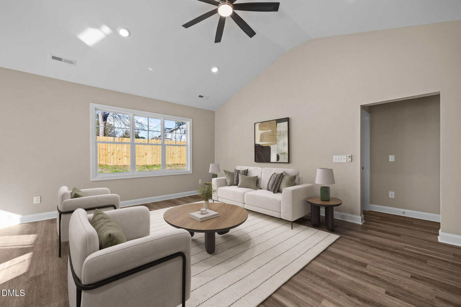 Modern living room with light walls, wood flooring, two armchairs, a sofa, round coffee table, side table, large windows, and a ceiling fan. Sunlight streams through the windows, brightening the space.