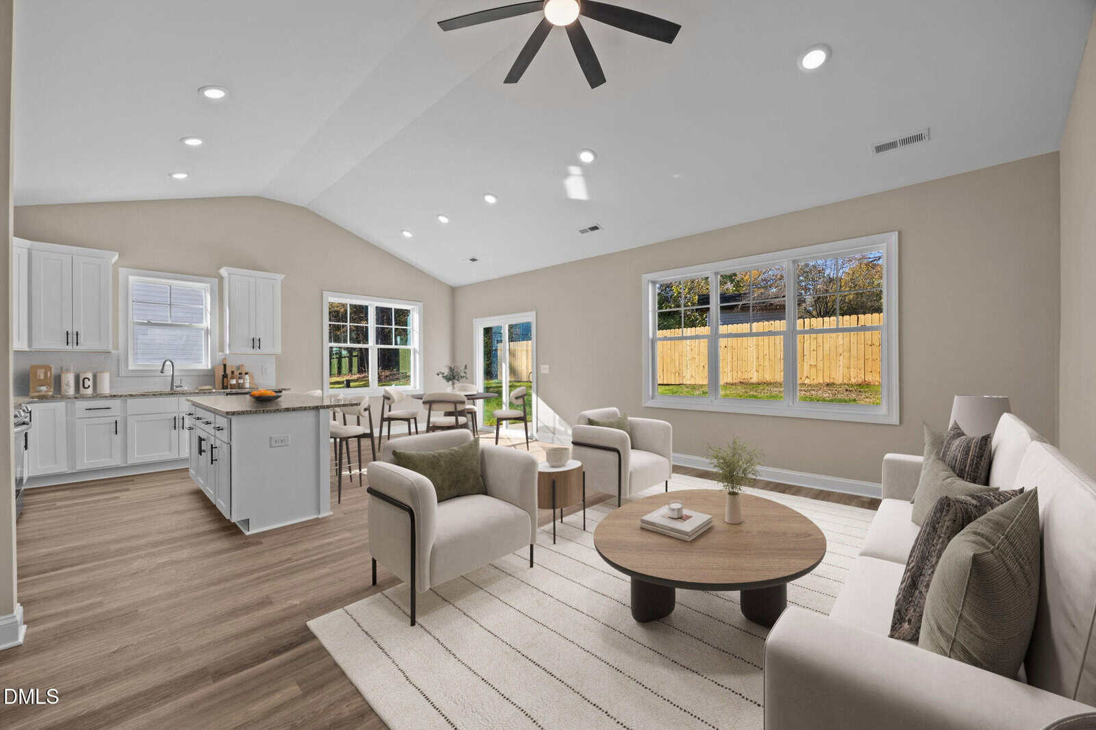 Bright, modern open-plan living room and kitchen with neutral décor, large windows providing outdoor views, light wood flooring, white cabinets, and a ceiling fan.