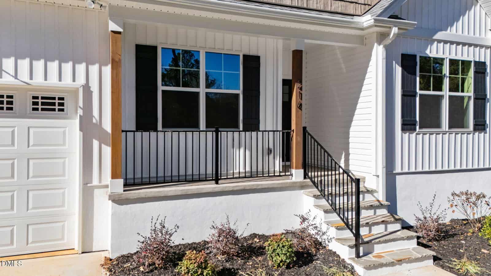 A small front porch with black metal railing, two wooden columns, and steps leading to a white house with black shutters and a garage door on the left. There are small shrubs in the garden bed below the porch.