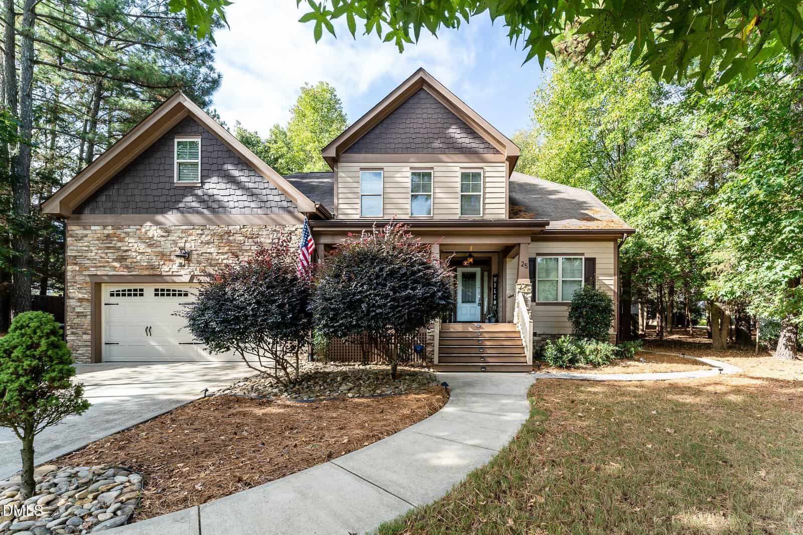 Two-story suburban house with stone and wood exterior, a covered front porch, and attached two-car garage. Concrete driveway curves toward the entrance, with landscaped shrubs and trees surrounding the lawn.