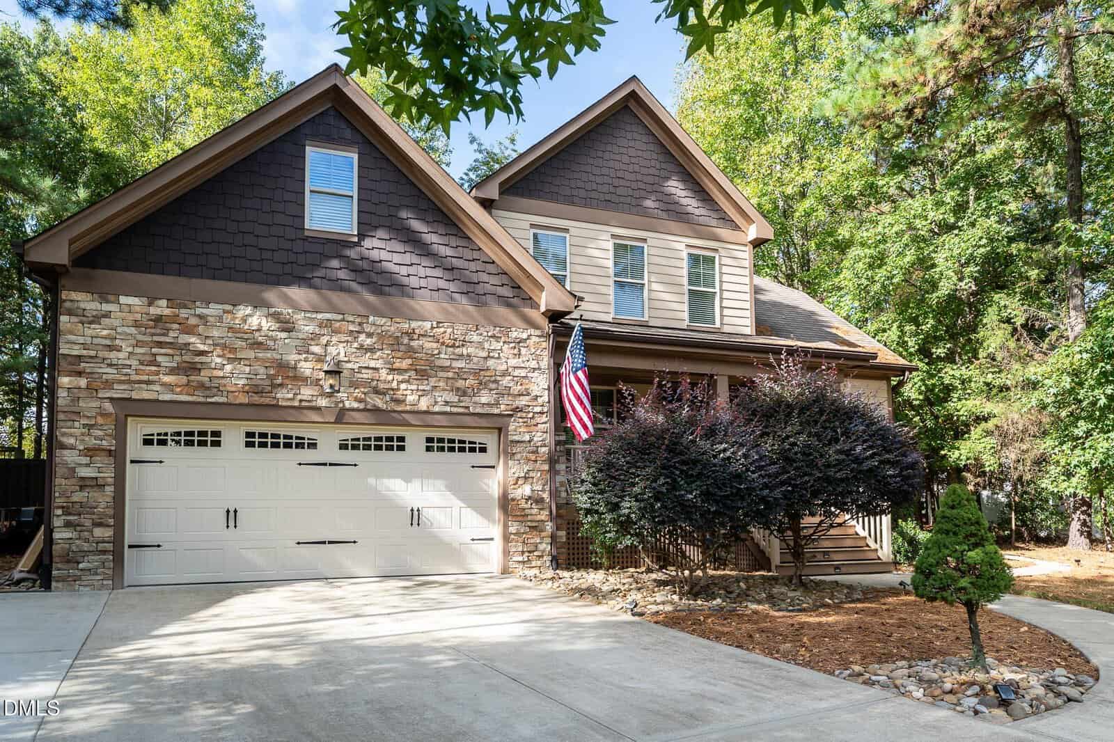 Two-story house with stone and brown siding exterior at 25 Paddy Lane, Youngsville. Features white double garage doors, an American flag by the porch, and a landscaped yard with trees and shrubs on a sunny day.