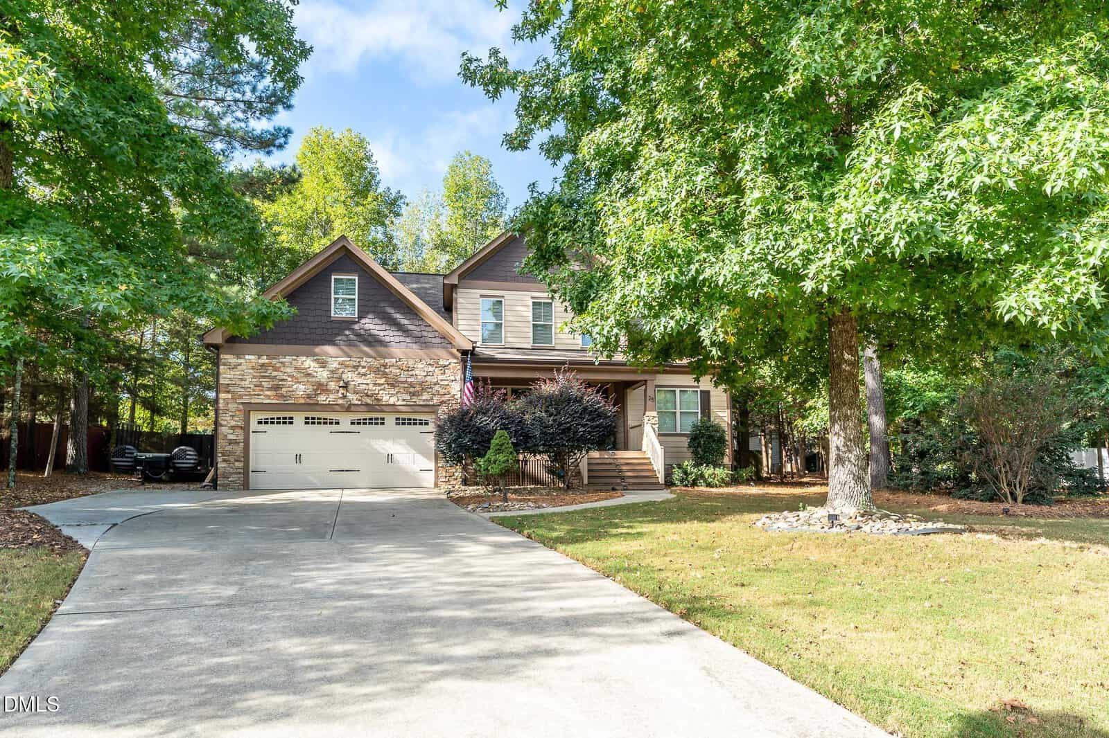 A two-story house with stone and brown siding, a two-car garage, front porch, and an American flag. The house is surrounded by trees and has a concrete driveway leading to the street.