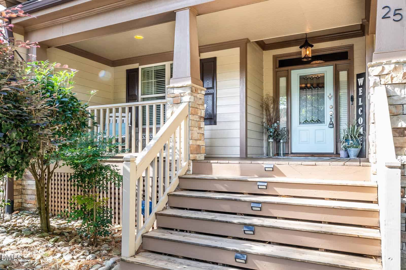 Front porch of a beige house with stone accents, white railing, wooden steps with built-in lights, a welcome sign, potted plants, and a glass front door with sidelights and a decorative wreath.