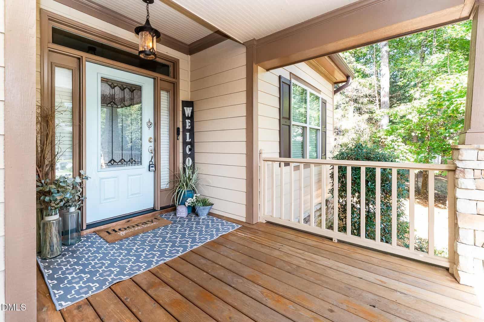 A covered wooden porch with a light blue front door, decorative glass panels, a patterned rug, potted plants, and a vertical "WELCOME" sign beside the door. Trees and greenery are visible in the background.