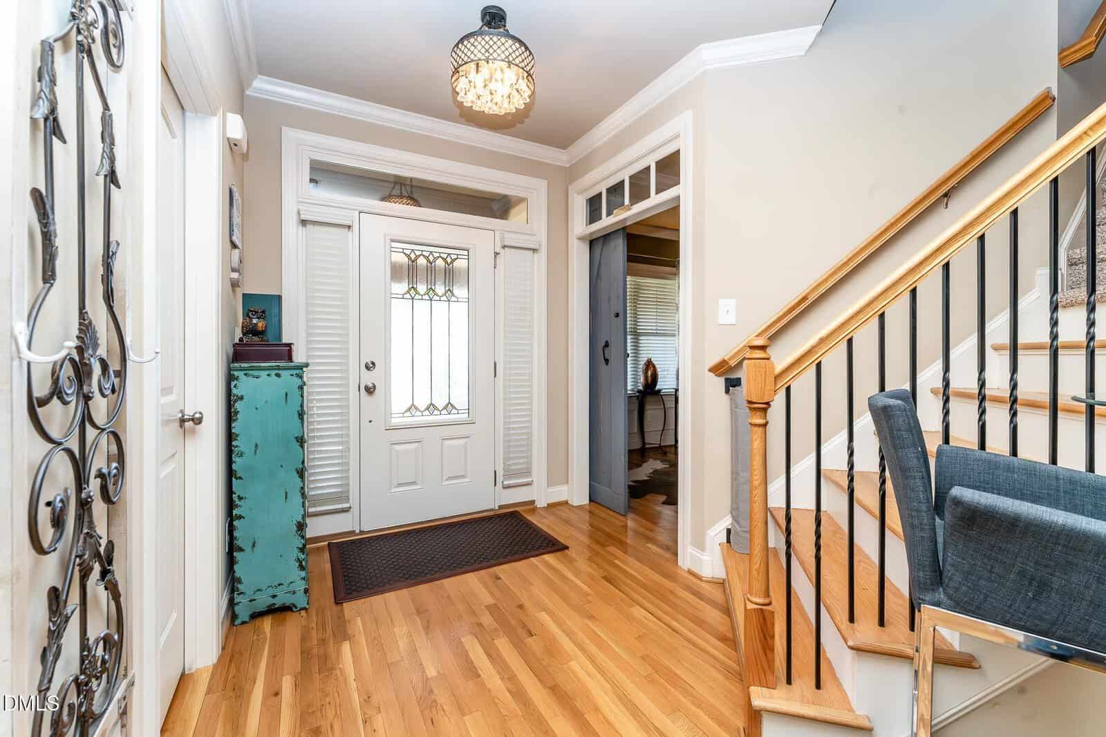 A bright entryway with light hardwood floors, a white front door with stained glass, sidelights and a transom window, a turquoise cabinet, and a staircase with wooden steps and black railings.