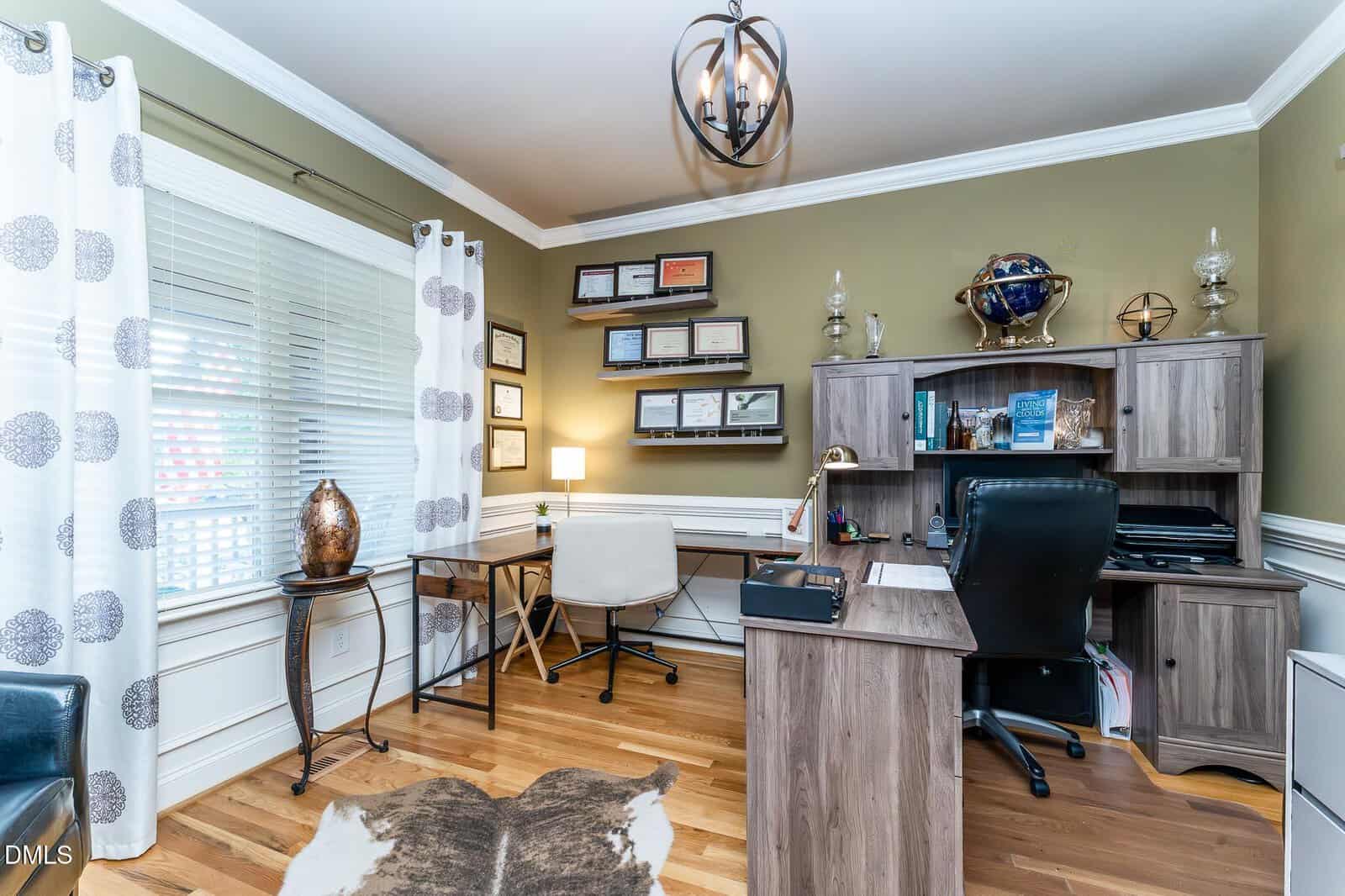 A home office with a wooden desk, shelving, a globe, and various certificates on the wall. A window with patterned curtains lets in light, and a white chair sits at a smaller desk by the window. Hardwood floor with a cowhide rug.