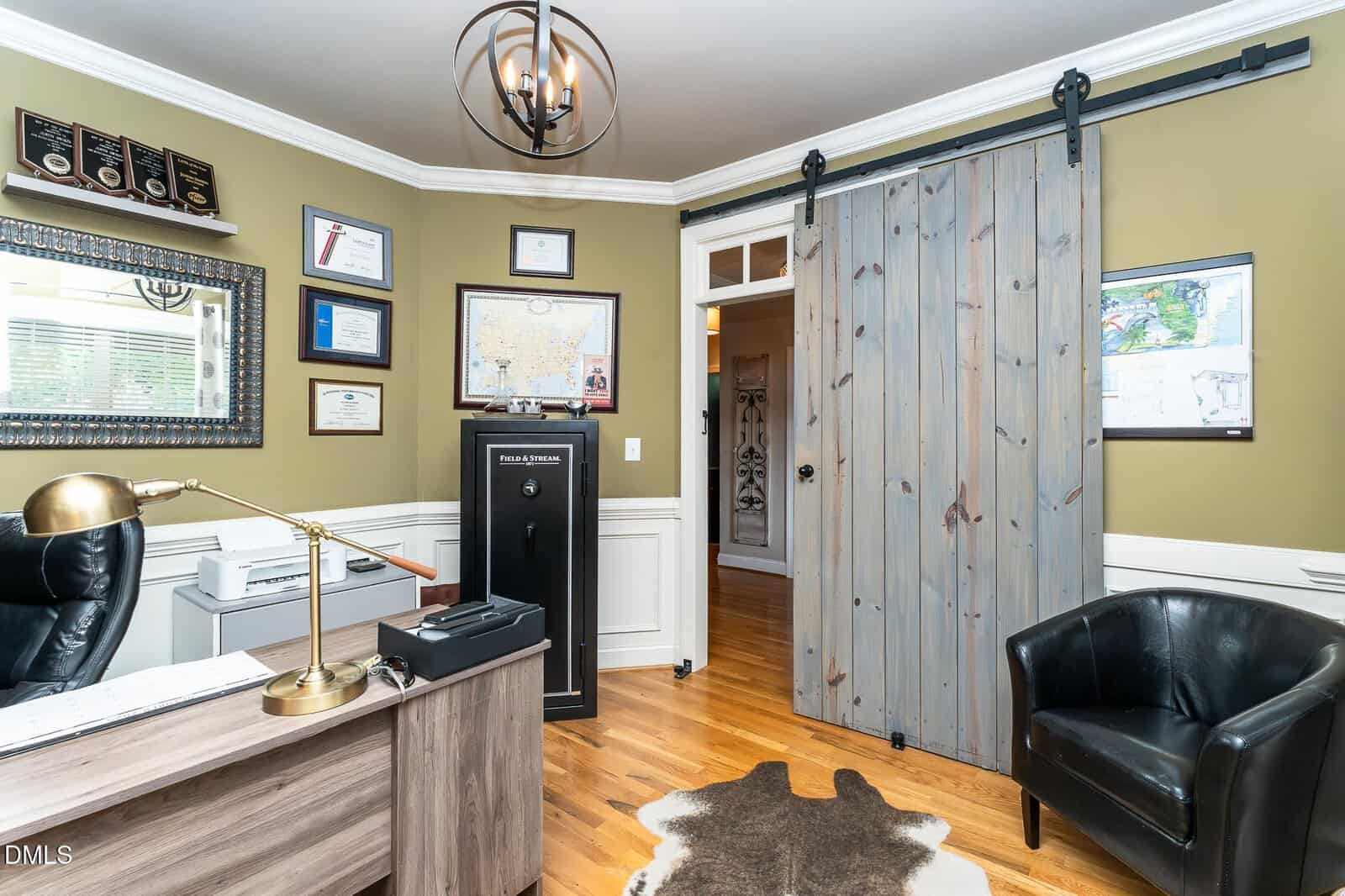 Home office with a desk, office chair, black armchair, cowhide rug, wall-mounted awards, framed certificates, a printer, a safe, and a sliding barn door partially open, revealing a hallway.