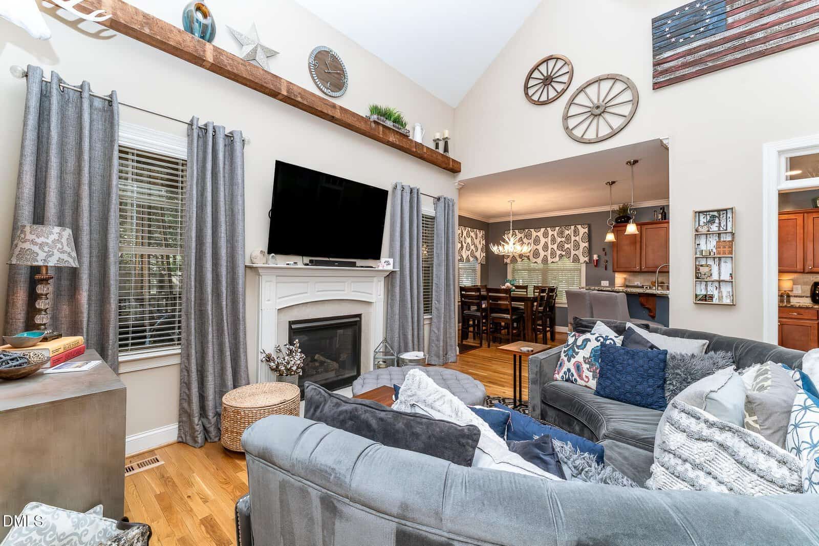 A living room with a gray sectional sofa, fireplace, wall-mounted TV, and large windows with gray curtains. The room opens to a dining area and kitchen, with various decorative items on the walls and shelves.