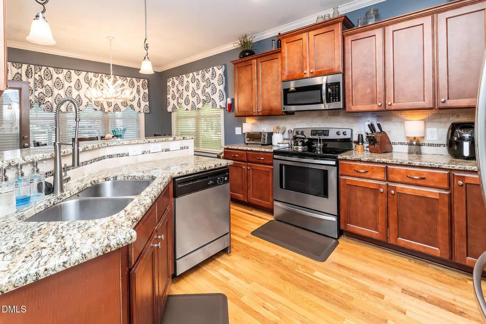 Modern kitchen with granite countertops, stainless steel appliances, wooden cabinets, a double sink, and hardwood floors. Pendant lights and patterned valances hang over windows in the background.