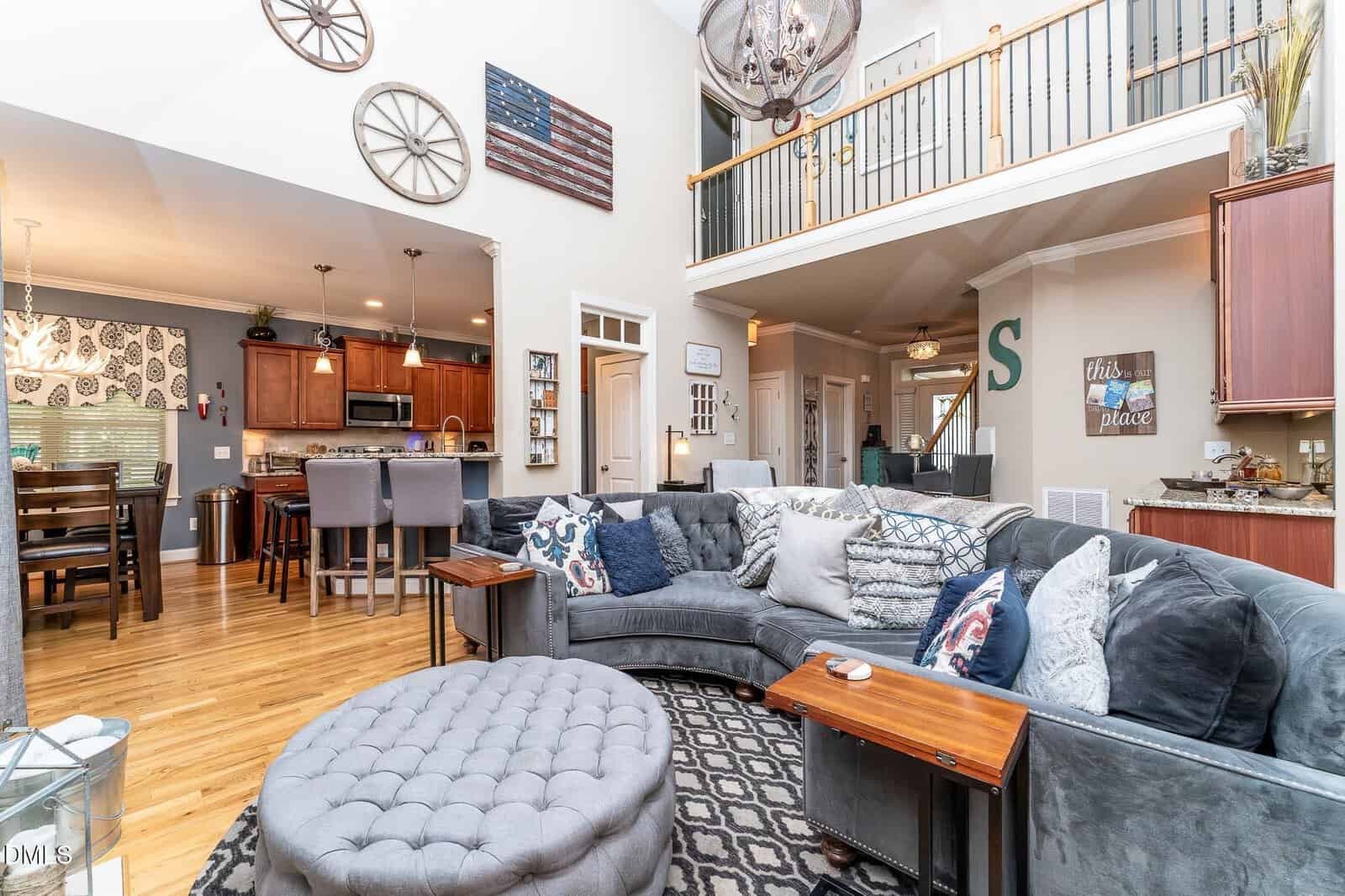 Spacious living room with gray sectional sofa, ottoman, and pillows, open to kitchen and dining area; wood floors, high ceiling, upstairs railing, and various wall decor, including an American flag and clocks.