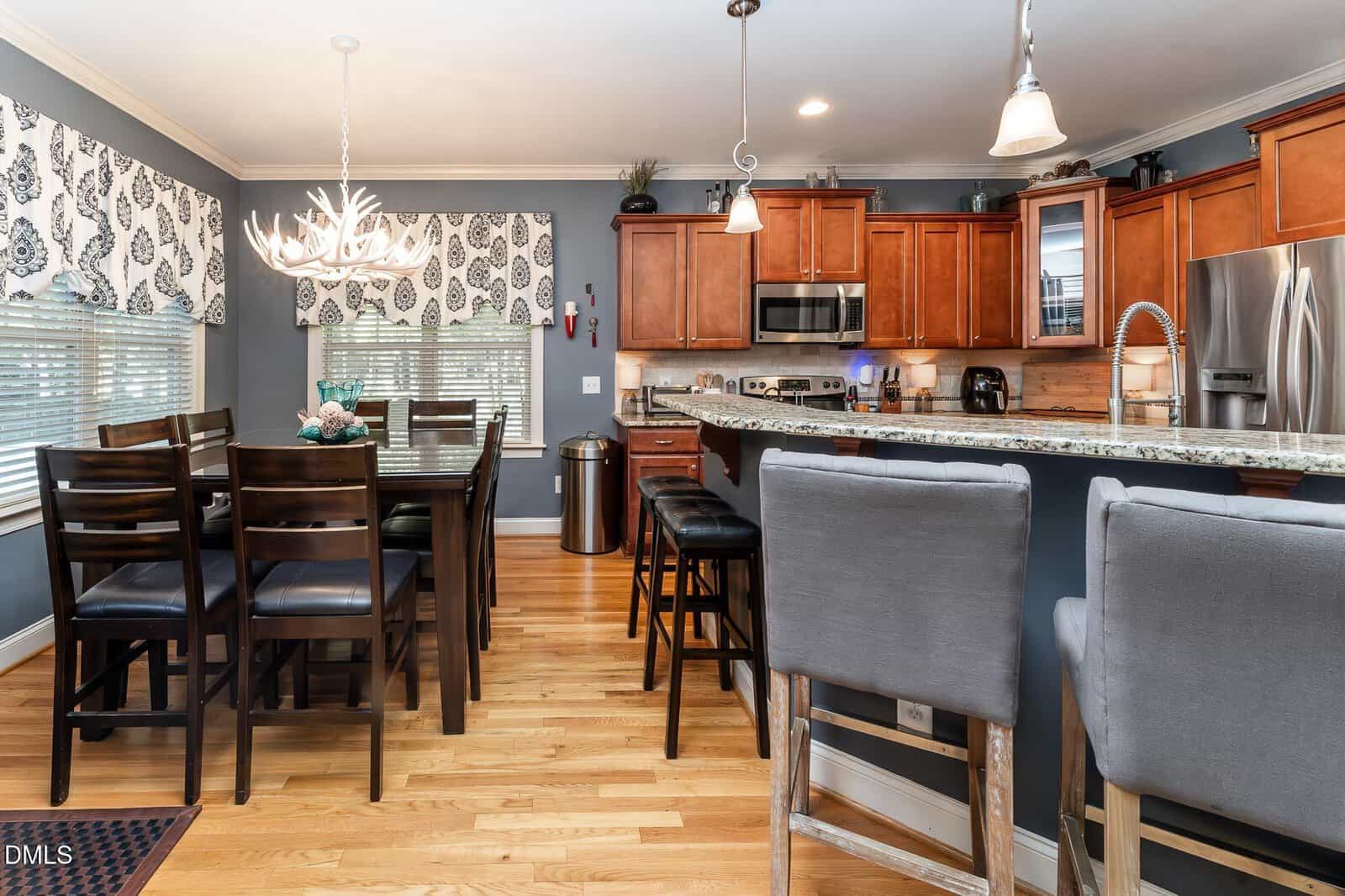 Modern kitchen with wooden cabinets, stainless steel appliances, granite countertops, a dining table with dark chairs, and two gray barstools at a kitchen island, all on light hardwood floors.
