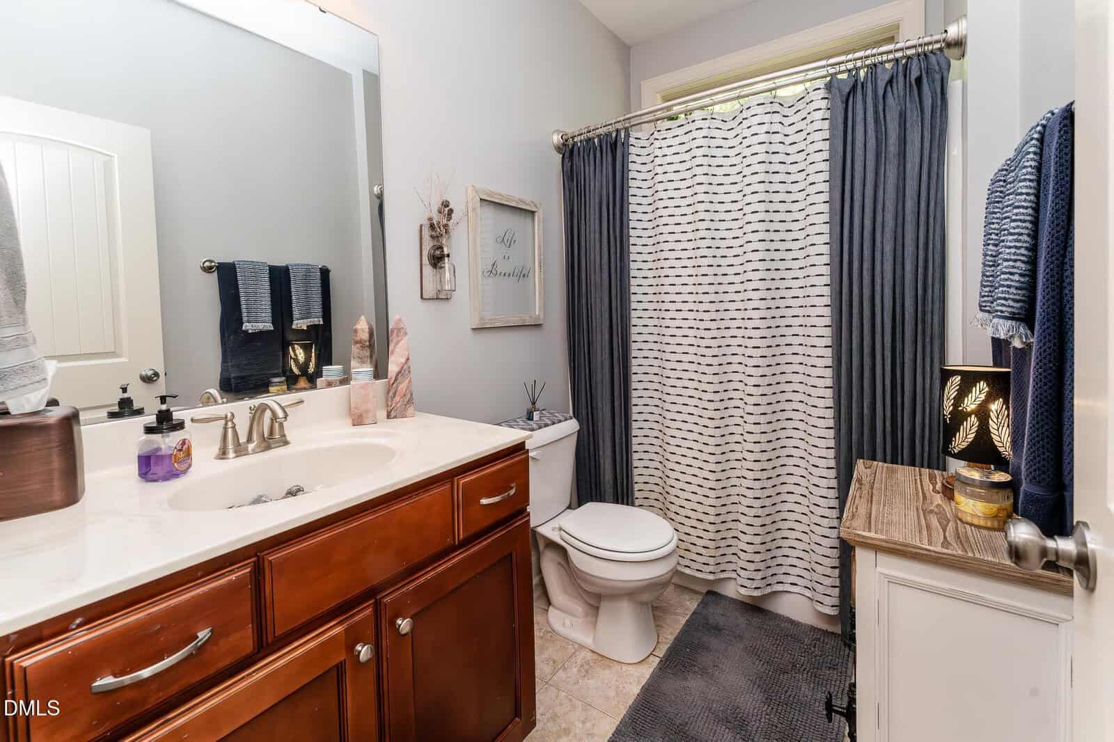 Bathroom with a wooden vanity, white sink, and marble countertop. Toilet next to a small cabinet with decor. Gray walls, a shower with a white and gray curtain, towel racks, and a wall sign complete the room.