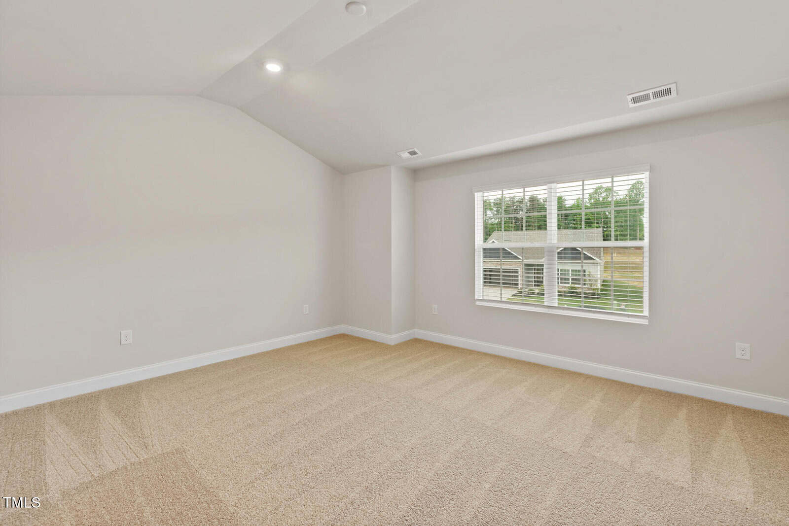 Empty, carpeted room with light beige walls, a vaulted ceiling with recessed lights, and a window with blinds letting in natural light; electrical outlets and vents are visible.
