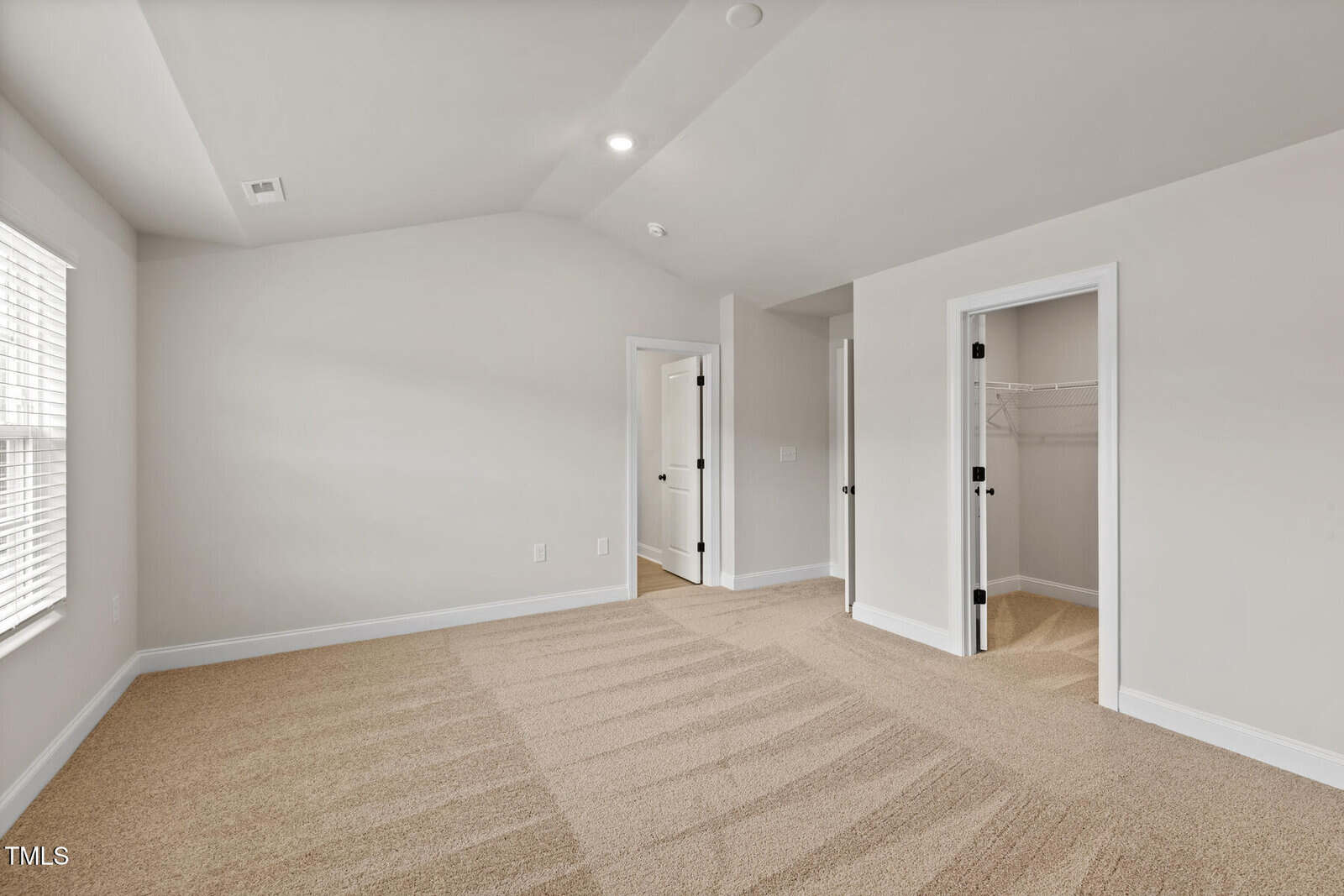 Empty, carpeted bedroom with beige walls, two windows on the left, and three doors: one open to a closet, one open to a hallway or bathroom, and one closed. Natural light fills the space.