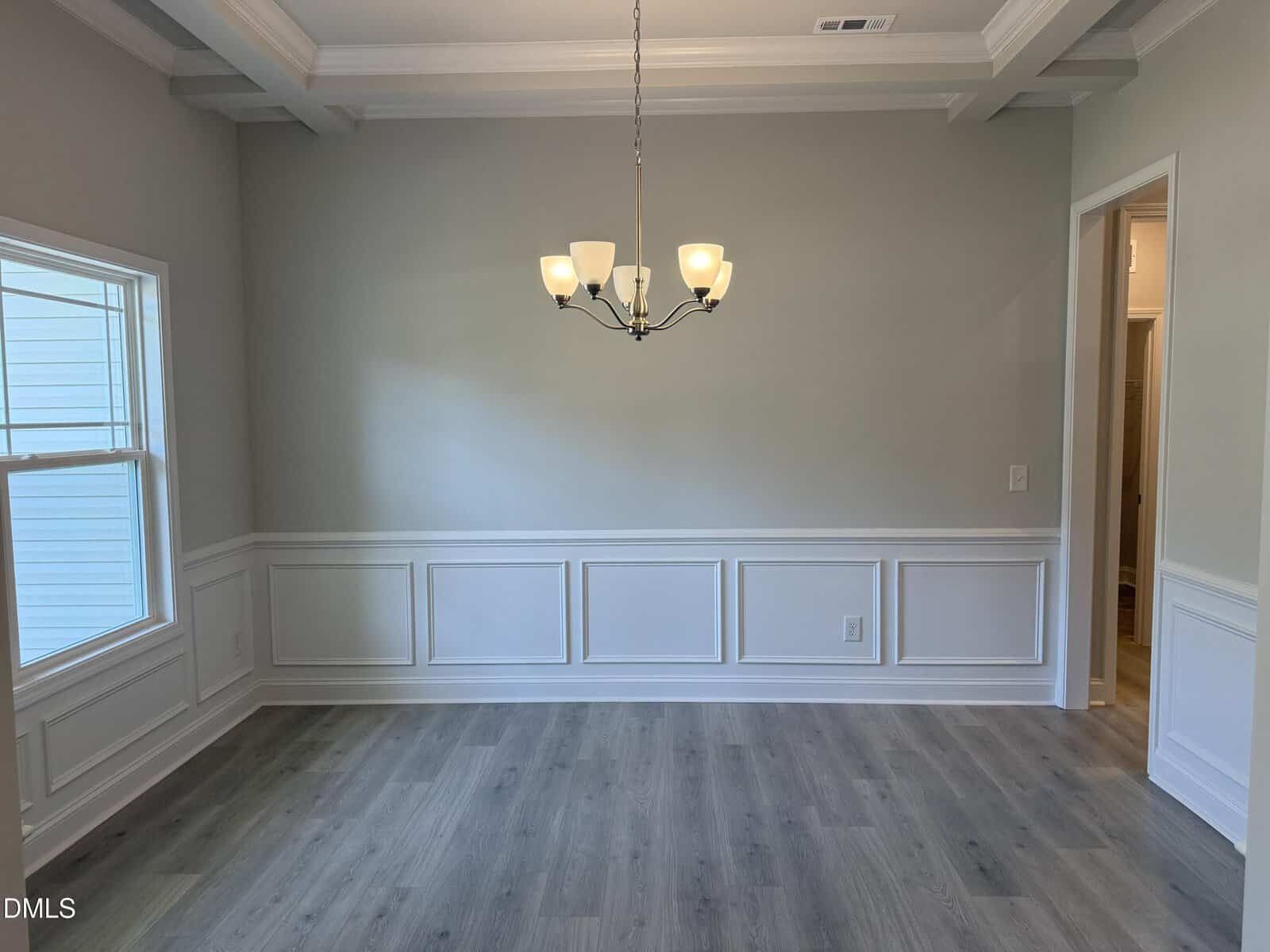 A bright, empty dining room with light gray walls, white wainscoting, gray wood floors, a window on the left, and a modern chandelier hanging from a coffered ceiling.