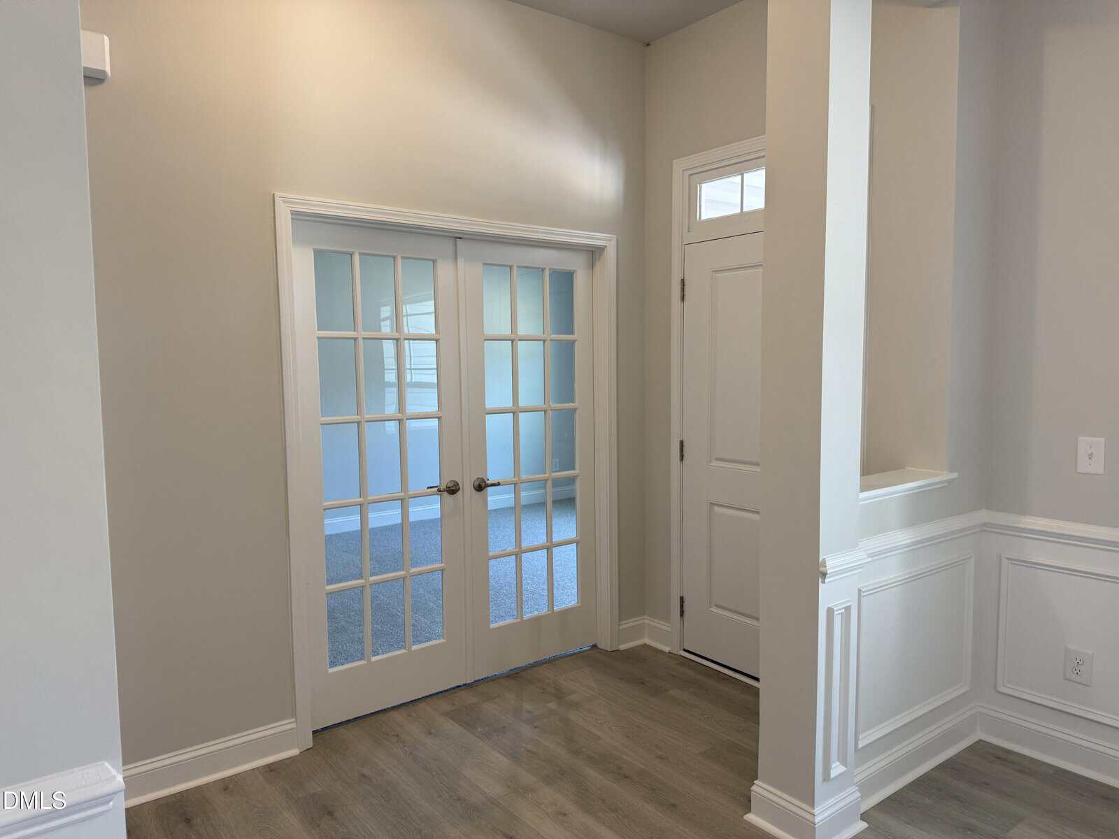 A bright entryway with light wood flooring, white paneled walls, and French doors with glass panes leading to another room. There is also a white front door and high ceilings.