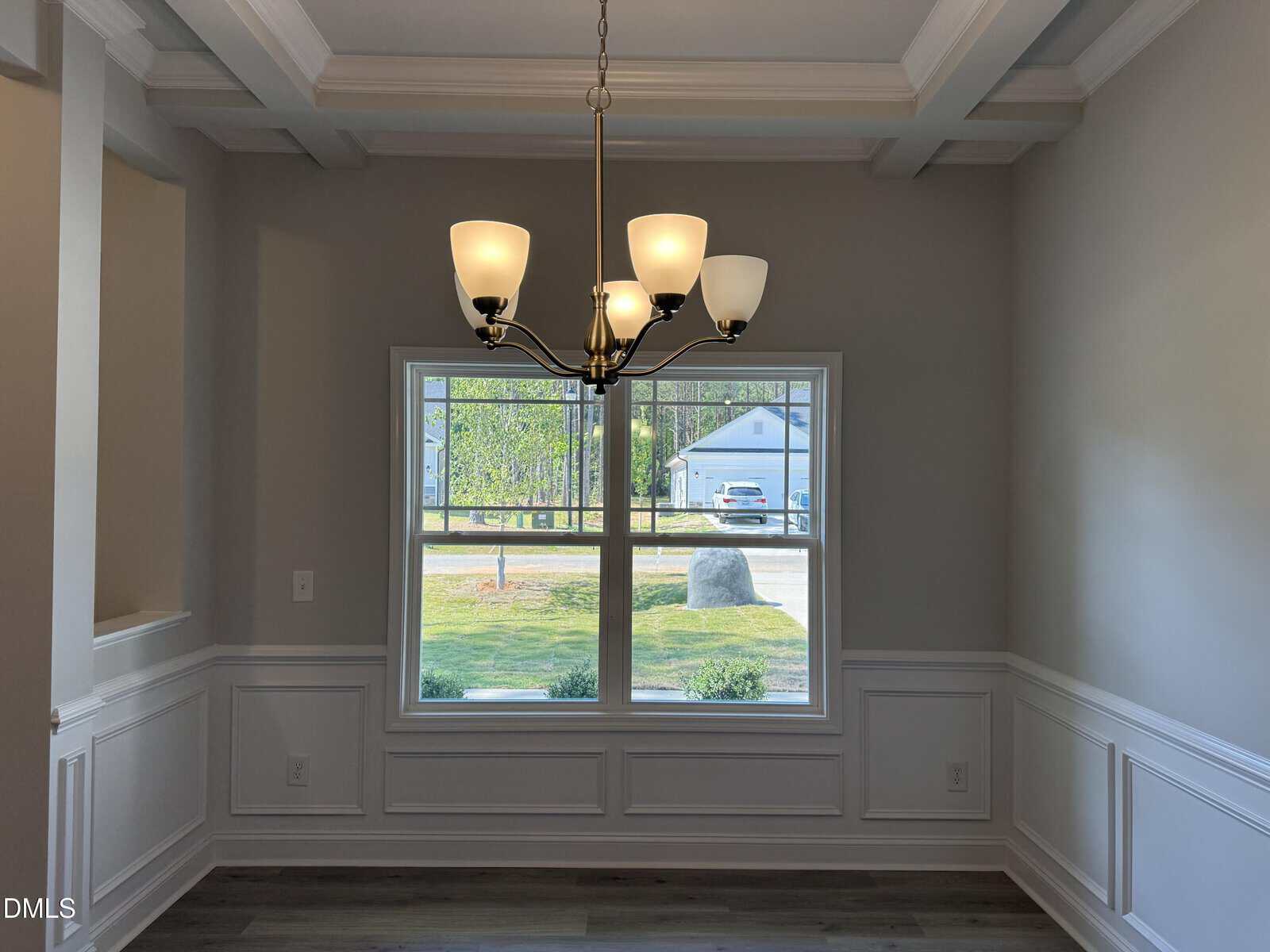A dining room with white wainscoting, gray walls, wood-style flooring, a modern brass chandelier, and a large window overlooking a yard with trees and a house in the background.