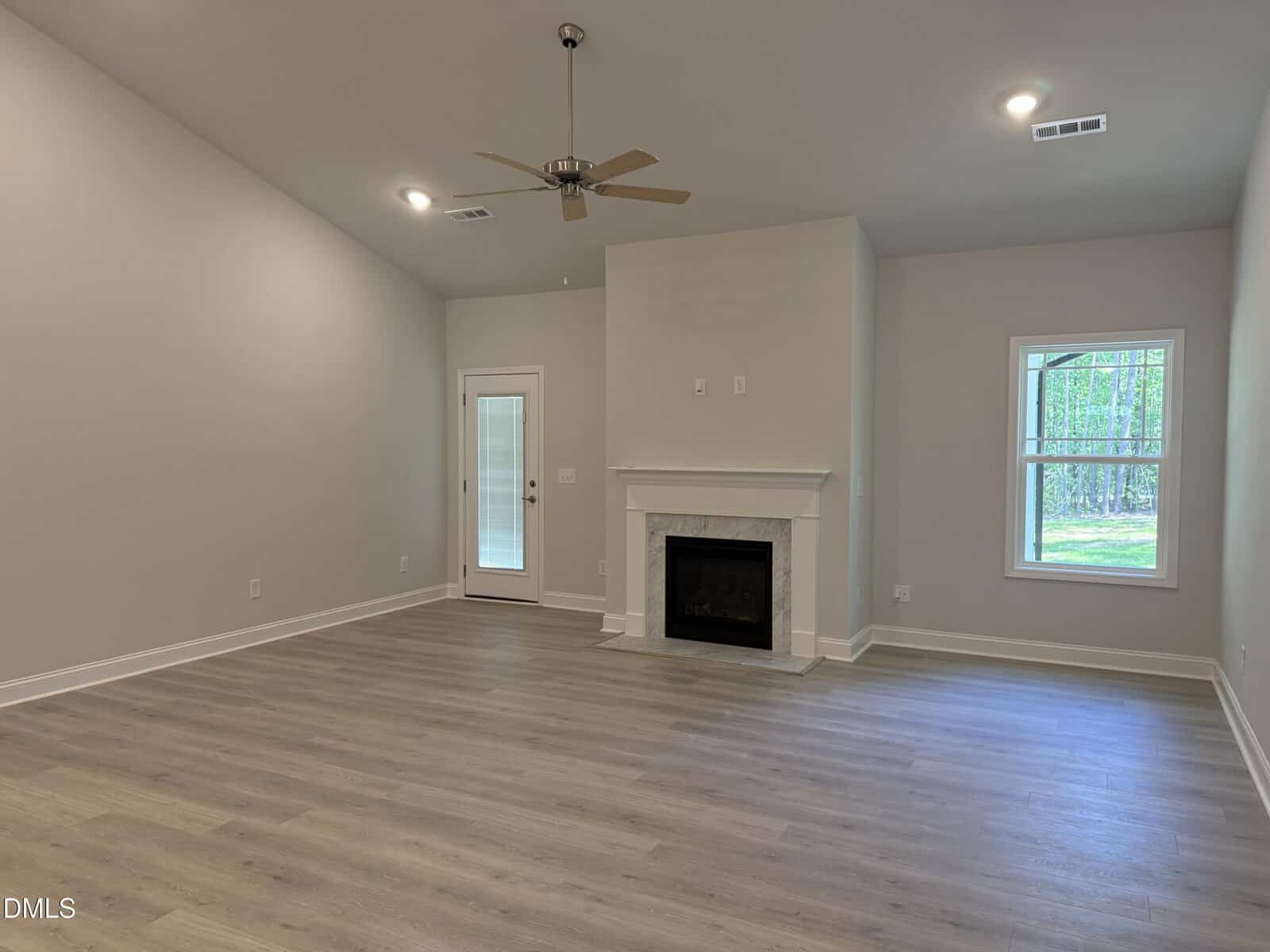 A spacious, empty living room with light wood flooring, a white fireplace, ceiling fan, recessed lighting, a window, and a glass door leading outside. Walls are light-colored, and the room has natural light from the window.