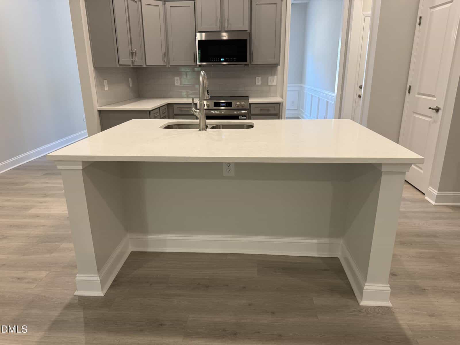 Modern kitchen with light wood floors, gray cabinets, white quartz island with sink and faucet, stainless steel appliances, and neutral walls.
